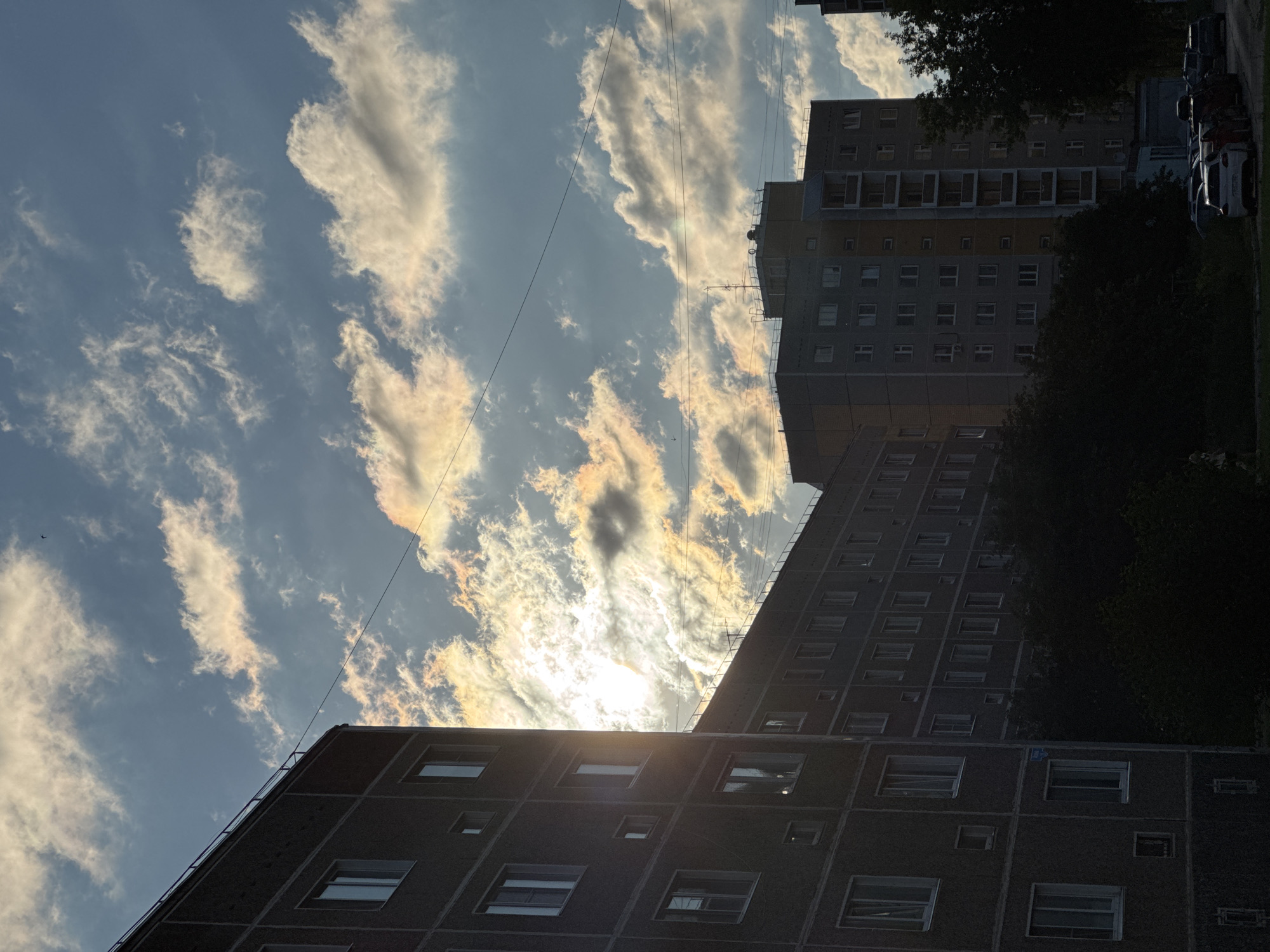 The image captures an urban scene at an angle looking upwards towards a bright, cloudy sky. In the foreground, to the left, is the side of a tall, dark-colored apartment building with visible windows. The building is silhouetted against the sky. To the right of this building, a second, lighter-colored apartment building is visible, also with many windows, extending further into the distance. A gap between these buildings reveals the bright sun partially obscured by clouds, creating a dramatic lighting effect with yellow and white hues. Thin, dark power lines are stretched across the sky. In the lower right portion of the image, the dark shapes of trees and the roofs of parked cars are visible, suggesting a street or parking area. The overall impression is one of an evening or late afternoon sky, with the setting or rising sun casting a warm glow on the clouds. The architecture suggests a Soviet-era residential block, commonly found in cities like Chaikovsky, Russia.