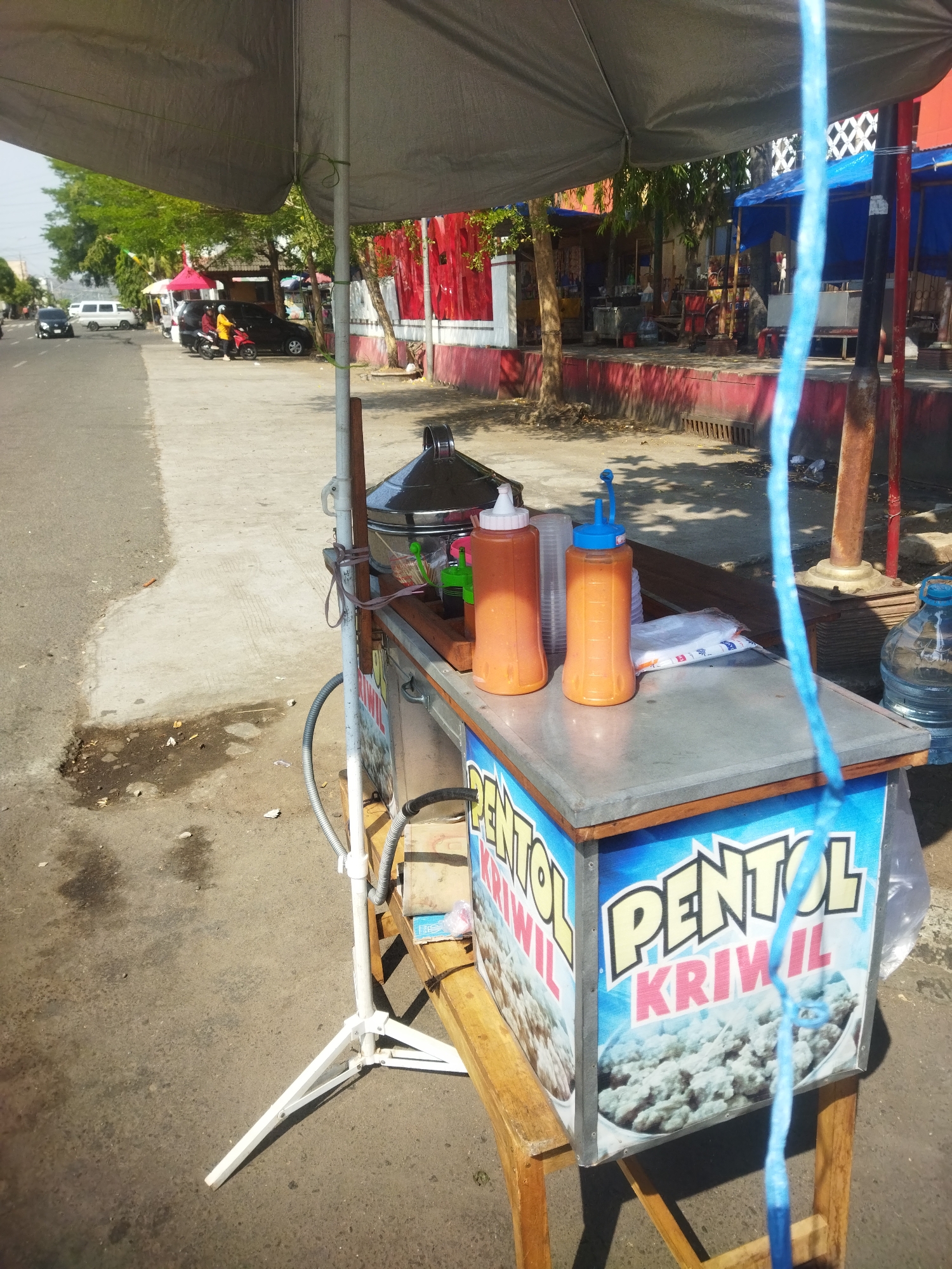An outdoor food cart, likely located on a roadside in Wonogiri, Indonesia, is depicted. The cart is covered by a large, gray umbrella. On the cart's surface, there are two orange squeeze bottles filled with sauce, plastic cups, and a large metal steaming pot. The front of the cart features a colorful graphic advertising "PENTOL KRIWIL" (a type of Indonesian snack) with an image of the food. A large blue plastic jug of water sits to the right of the cart. The background shows a street with some vehicles, trees, and buildings, suggesting an urban or semi-urban setting. The ground is paved asphalt with some debris. A blue rope hangs vertically in the foreground, obscuring part of the cart. The scene appears to be a sunny day, with shadows cast on the ground.