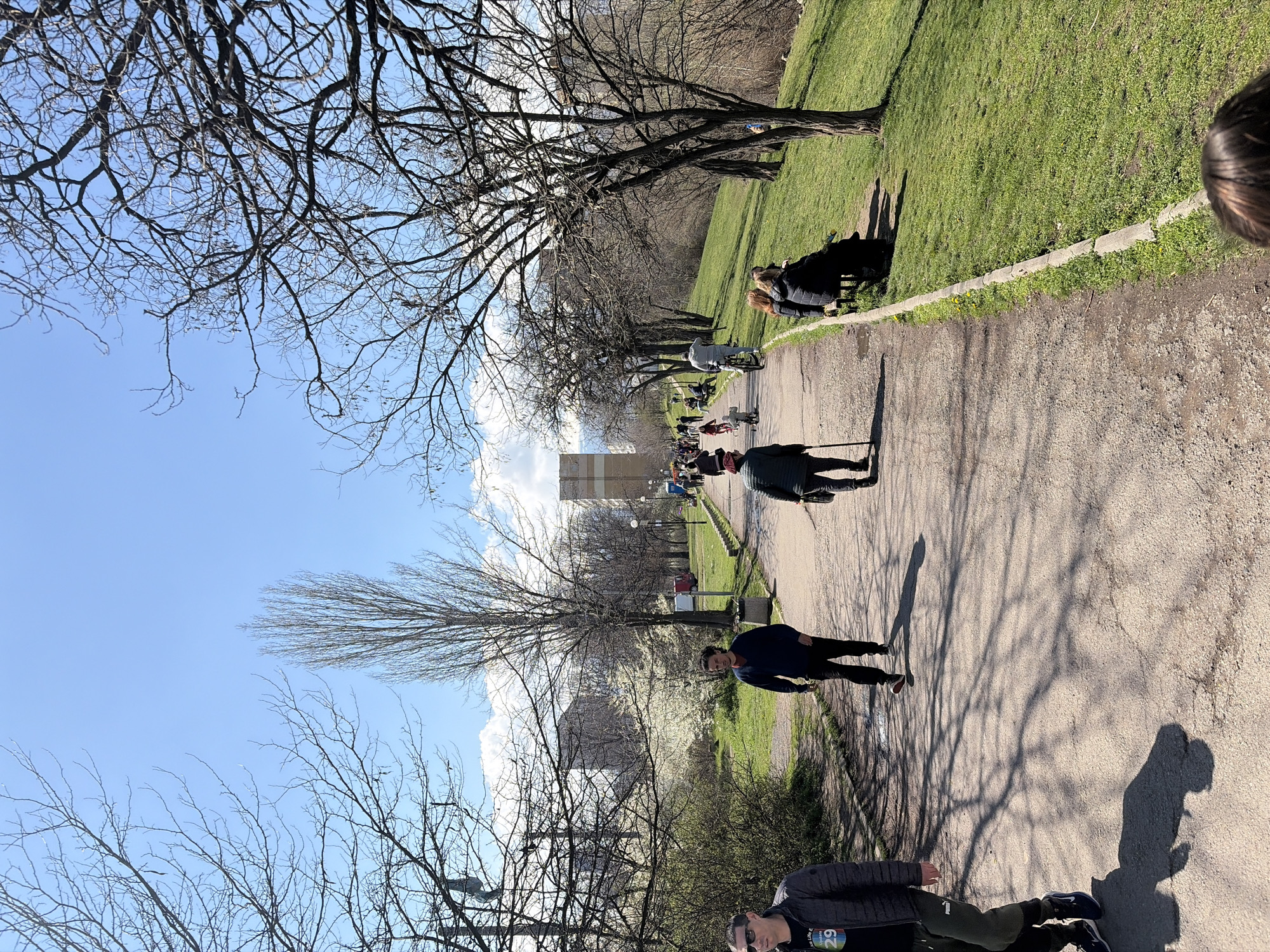 The image shows a park pathway on a sunny day, likely in early spring. A number of people are visible, some walking, others sitting on benches or strolling. A few bare trees are lining the pathway, casting shadows on the ground. In the background, a building and blooming trees are visible, indicating a city environment. Based on the style of the architecture and the overall feel of the environment, it is likely in Sofia, Bulgaria, although no specific landmarks are visible to confirm this definitively. The composition of the image is from a lower angle.