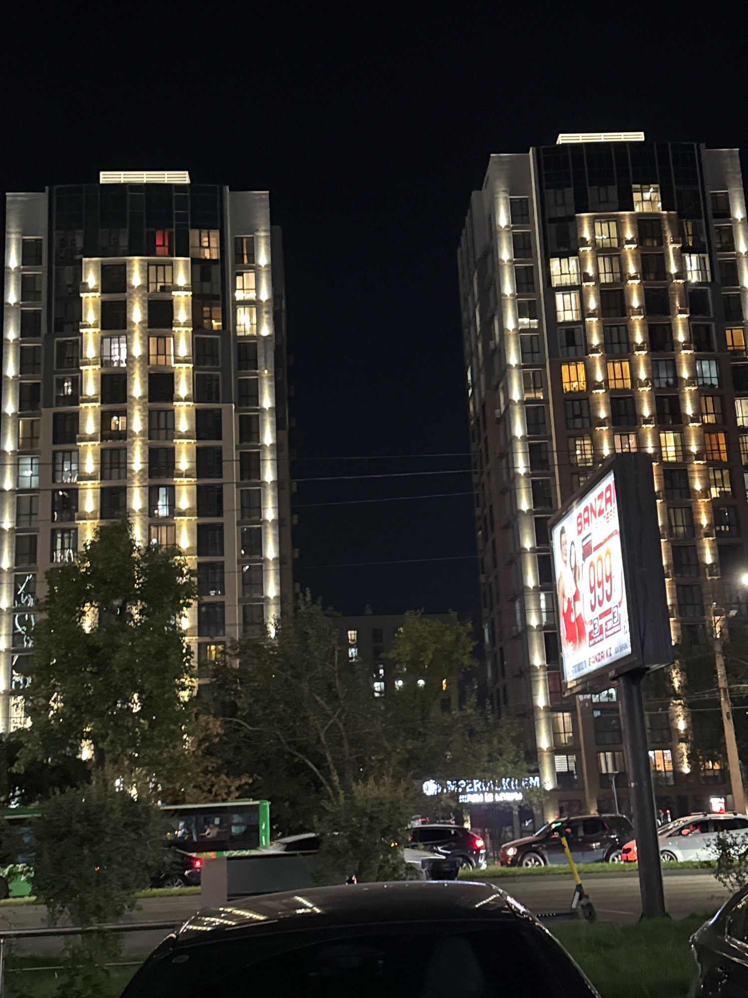 This is a nighttime, eye-level, outdoor photograph taken in Almaty, Kazakhstan. The scene is dominated by two modern, illuminated high-rise apartment buildings, one on the left and one on the right of the frame, facing each other across a street. The buildings are adorned with vertical bands of warm, yellow exterior lighting, and many windows are lit from within, casting a glow into the dark sky.
On the right side of the frame, a large, illuminated billboard displays an advertisement. The advertisement features images of two women in red dresses and large red numerals reading "999". The text "BANZA" is visible at the top of the billboard. Below the buildings, trees with dark green foliage are visible. A street runs horizontally across the foreground, with several cars parked and driving on it. On the left side of the street, a green bus is visible. The ground appears to be a paved road, with some grassy areas and sidewalks. The overall atmosphere is urban and modern, with the lights of the buildings and the billboard creating a vibrant nighttime scene.