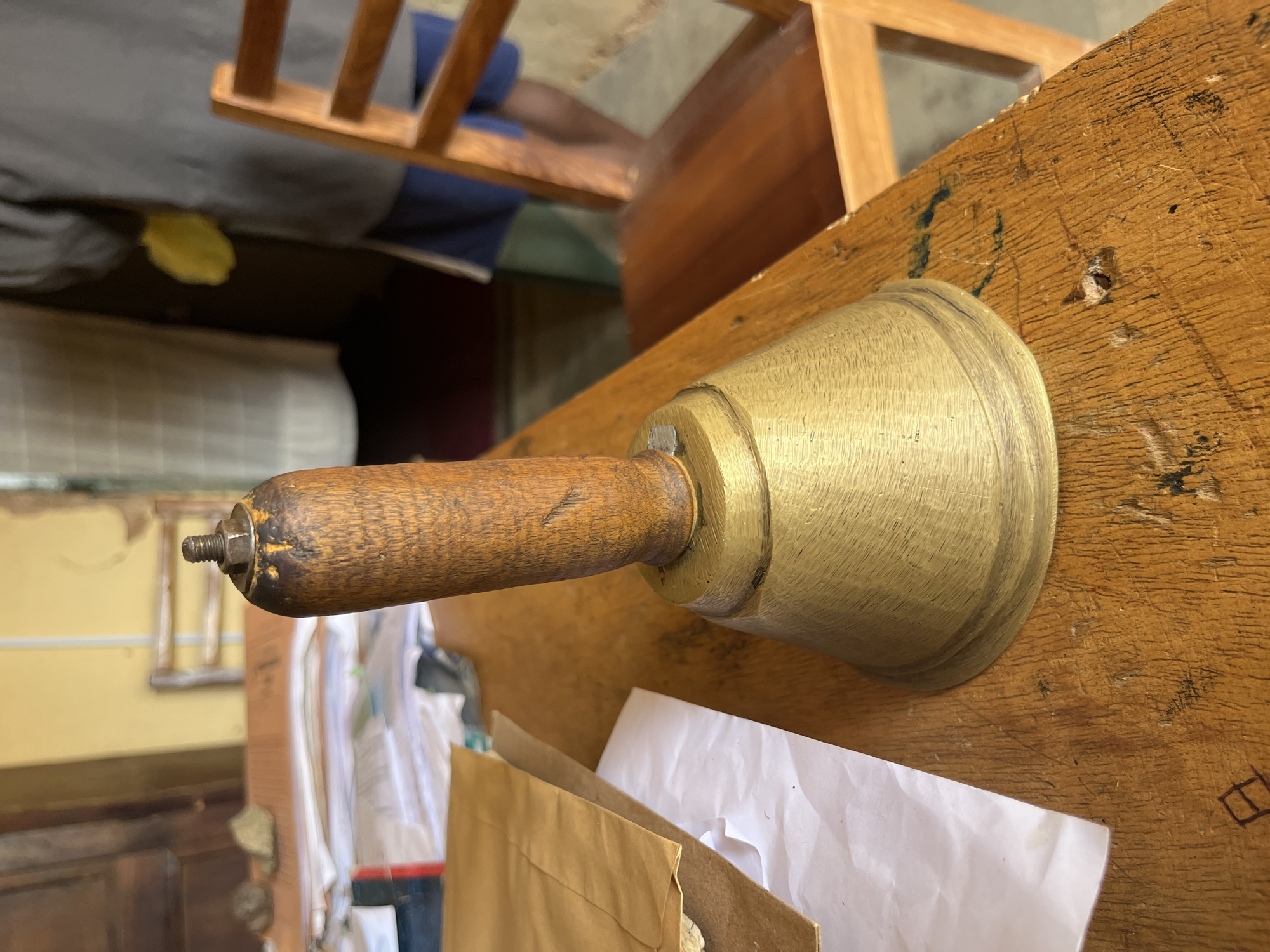 A close-up, low-angle shot shows an old brass bell with a wooden handle resting on a worn wooden desk. The bell's handle, made of dark wood, is capped with a metal screw. The bell itself is a classic conical shape, showing signs of age and use with a patina of discoloration. The desk surface is rough and textured, with visible scratches and dark marks, suggesting it is old and well-used.
Scattered on the desk are a few sheets of white paper and a brown paper envelope, suggesting a workspace or office environment. In the background, out of focus, is a wooden chair with a light wooden frame and slats. Behind that, further into the background, a person in a grey shirt and blue shorts is visible, holding a yellow object. The overall setting appears to be indoors, likely in a rustic or utilitarian building, possibly an office or classroom. The image is set in Bukuru, Nigeria, implying a local context for the scene.