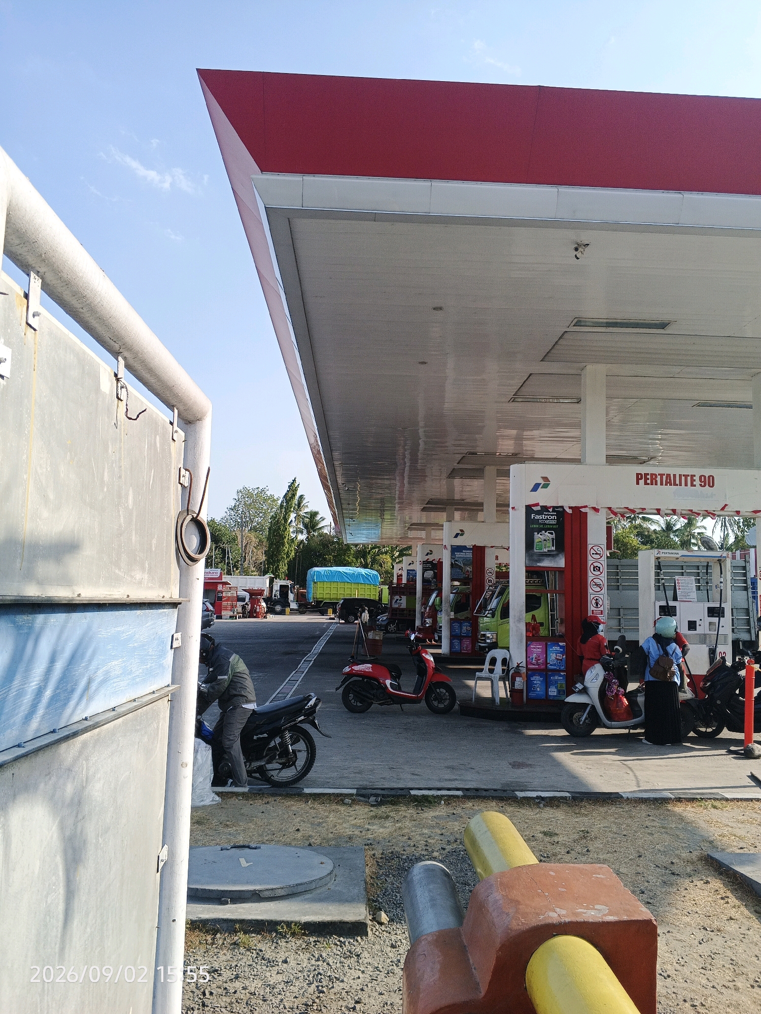 This is a medium shot, eye-level, outdoor photograph taken at a Pertamina gas station in Pinrang, Indonesia, on September 2, 2026, at 15:55. The image captures a bright, sunny day with a clear blue sky. In the foreground, a textured concrete and colorful pipe structure is visible. To the left, a gray metal wall with blue paneling partially obscures the view.
The main focus is the gas station canopy, characterized by its red roofline and white ceiling with visible lighting fixtures. Several red and white striped poles support the canopy. A prominent sign reads "PERTALITE 90".
Several people and vehicles are present. On the left side of the image, a person in a grey jacket and helmet is sitting on a black motorcycle, appearing to be refueling or preparing to leave. In the mid-ground, two red scooters are parked. Further to the right, a white scooter is parked near a gas pump. A woman in a blue top and light blue headscarf is standing next to the white scooter, and another woman in a red top and dark headscarf is attending to a gas pump. Behind them, a light green truck with a blue tarp-covered cargo is parked. Another truck, white with a red cab, is visible further in the background.
The ground is paved with asphalt and gravel. A circular manhole cover is set in concrete on the left side of the frame. The environment appears to be a typical roadside gas station with surrounding trees and tropical vegetation in the distance.