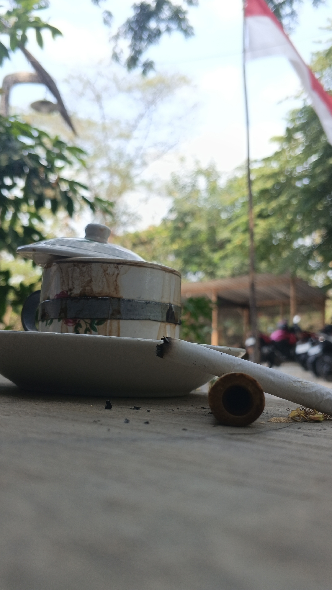 This is a close-up, low-angle outdoor photograph of a cigarette resting on a saucer next to a lidded cup. The cup appears to be ceramic with a floral pattern and decorative bands, and it has staining around the rim. Ash and small debris are scattered on the saucer and the wooden surface the objects are placed on. The cigarette is partially smoked and lies diagonally across the saucer, with its filter end facing the viewer. In the background, out of focus, are trees, a rustic shelter structure, and several motorcycles. A portion of a red and white Indonesian flag is visible in the upper right corner of the frame. The lighting suggests it is daytime. The scene evokes a sense of casualness and potentially a brief pause, possibly in a public space or outdoor seating area, given the motorcycles and shelter. Ngampel, Indonesia is mentioned as the location, suggesting a specific cultural or geographical context for this scene.