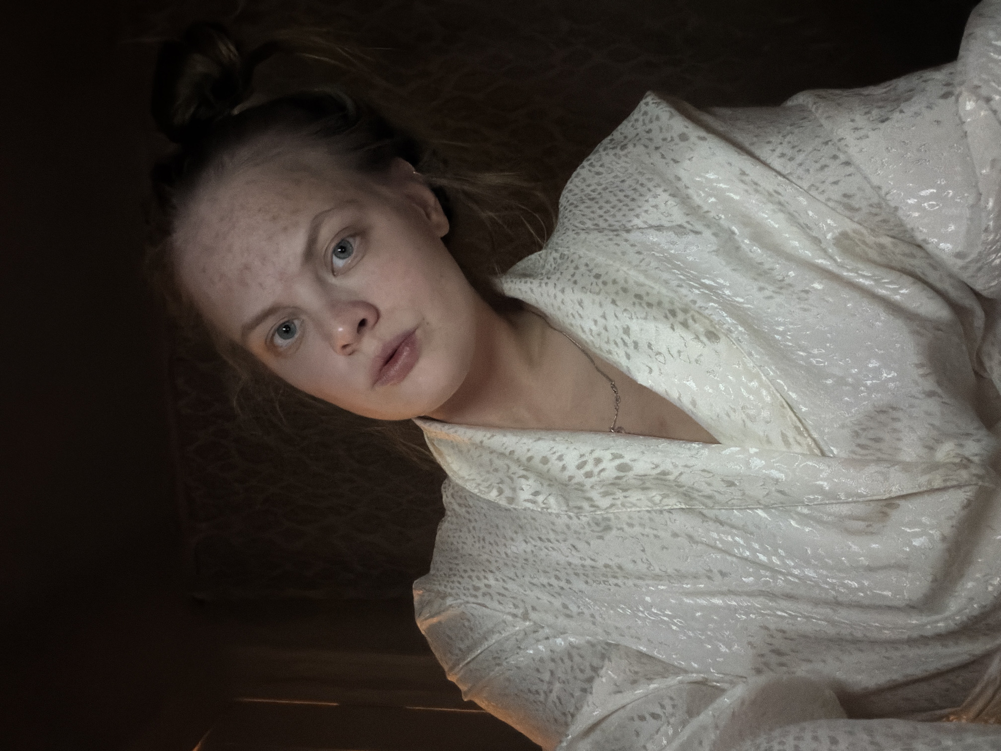 A medium shot depicts a woman with fair skin and blue eyes, who appears to be lying down. She is facing towards the camera. Her hair is pulled up in a bun on the top of her head. She is wearing a patterned, white robe. A thin silver necklace is visible on her neck. The background is dark and out of focus, but appears to be a room. The location is unknown and there is no information to suggest this photo was taken in Izhevsk, Russia.