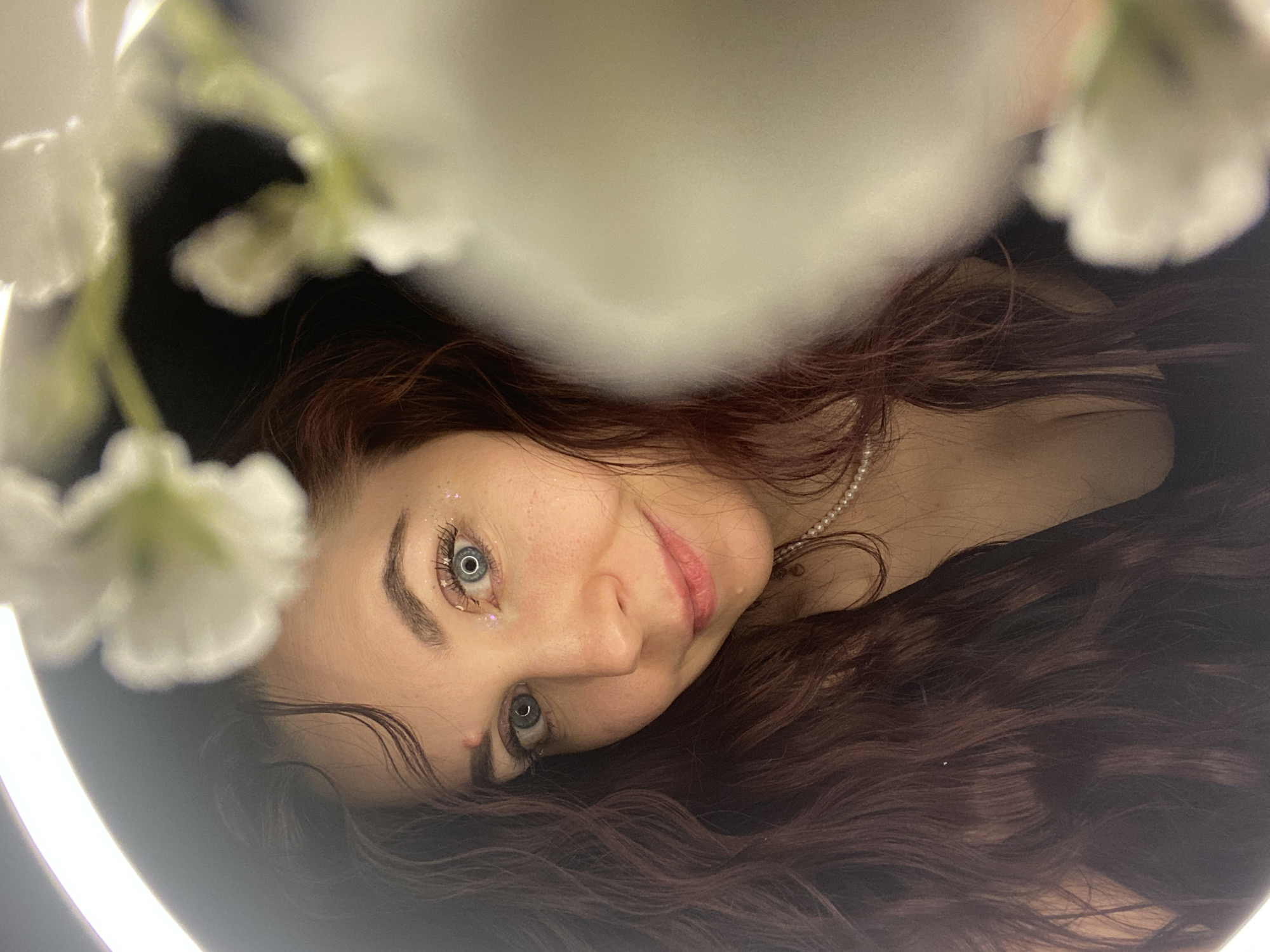 The image is a close-up, angled portrait of a fair-skinned woman with auburn hair. She has blue eyes, and sparkly makeup is visible around one eye. A pearl necklace is around her neck. White flowers are in the foreground, partially obscuring the view. The lighting suggests an artificial source, possibly a ring light, is being used. There is no information to suggest that the image was taken in the city of Chaikovsky, Russia.