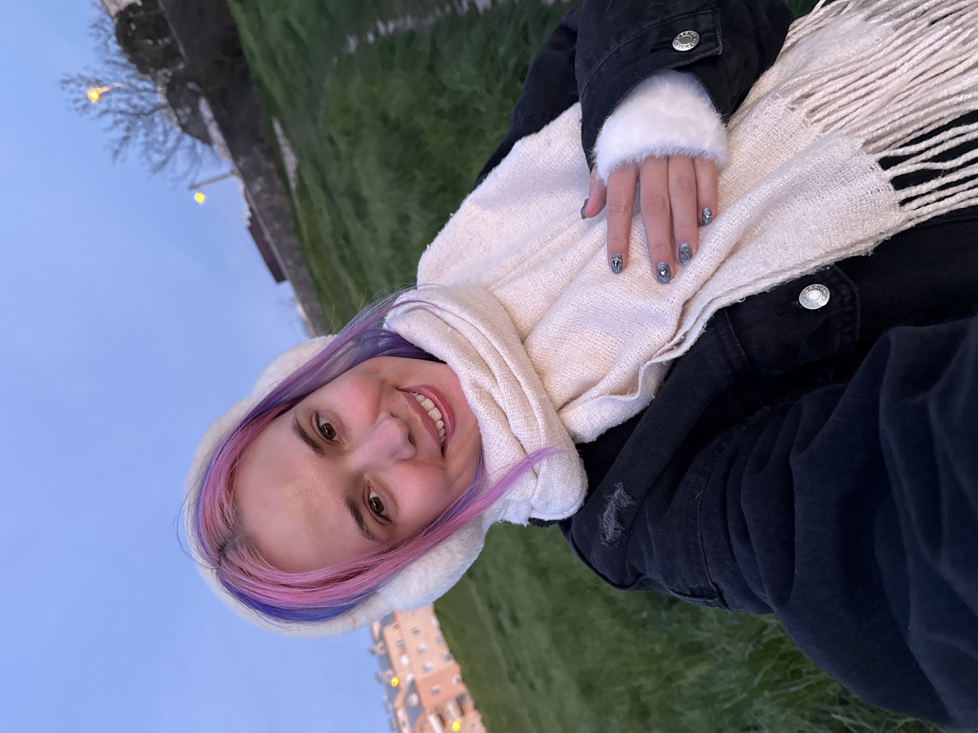 A woman with pink and purple hair is smiling and looking at the camera. She is wearing a white scarf and a black jacket with white fur cuffs. Her nails are decorated with silver designs. The background includes green grass, a building with lights, and a blue sky. The photo appears to be taken outdoors, possibly in the City of Zagreb, Croatia.