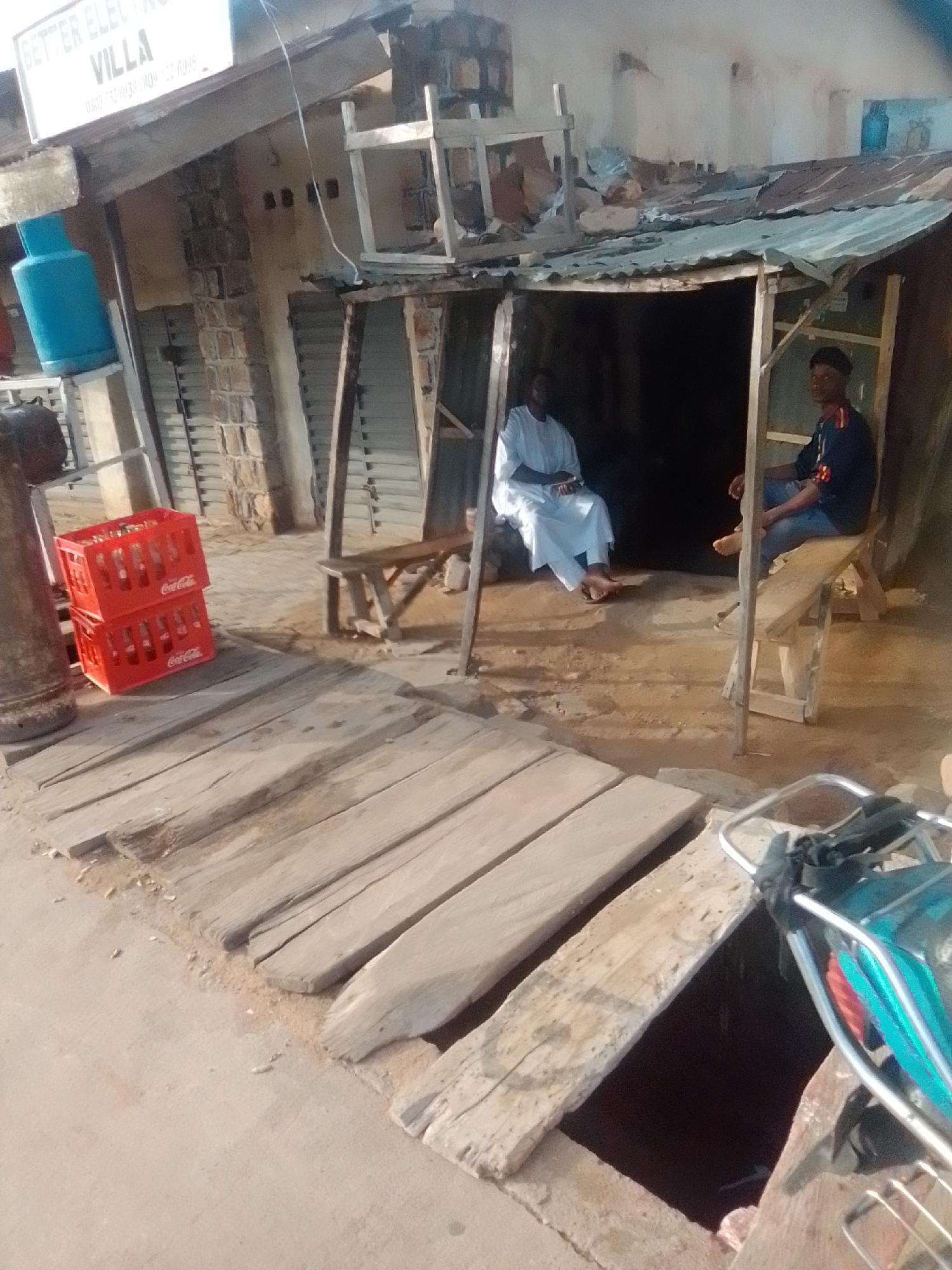The image depicts an outdoor scene in Pantame, Nigeria, featuring a small, makeshift business or dwelling. In the foreground, a wooden walkway with gaps is visible, leading towards a structure with a corrugated metal roof. To the left, two stacked red crates of Coca-Cola bottles are prominent. A blue gas cylinder stands on a metal rack near some stone-like structures and a shuttered opening.
Inside the structure, two men are seated. One man, wearing a white garment, is seated on the ground or a low surface. Another man, in a dark blue top and jeans, sits on a wooden bench. The environment appears rustic, with exposed building materials and dirt flooring. A trolley or cart with a blue and red object is partially visible on the right side of the image, suggesting a commercial or transport-related activity. A sign with "BETTER ELECTRIC VILLA" and a phone number is mounted above the structure.