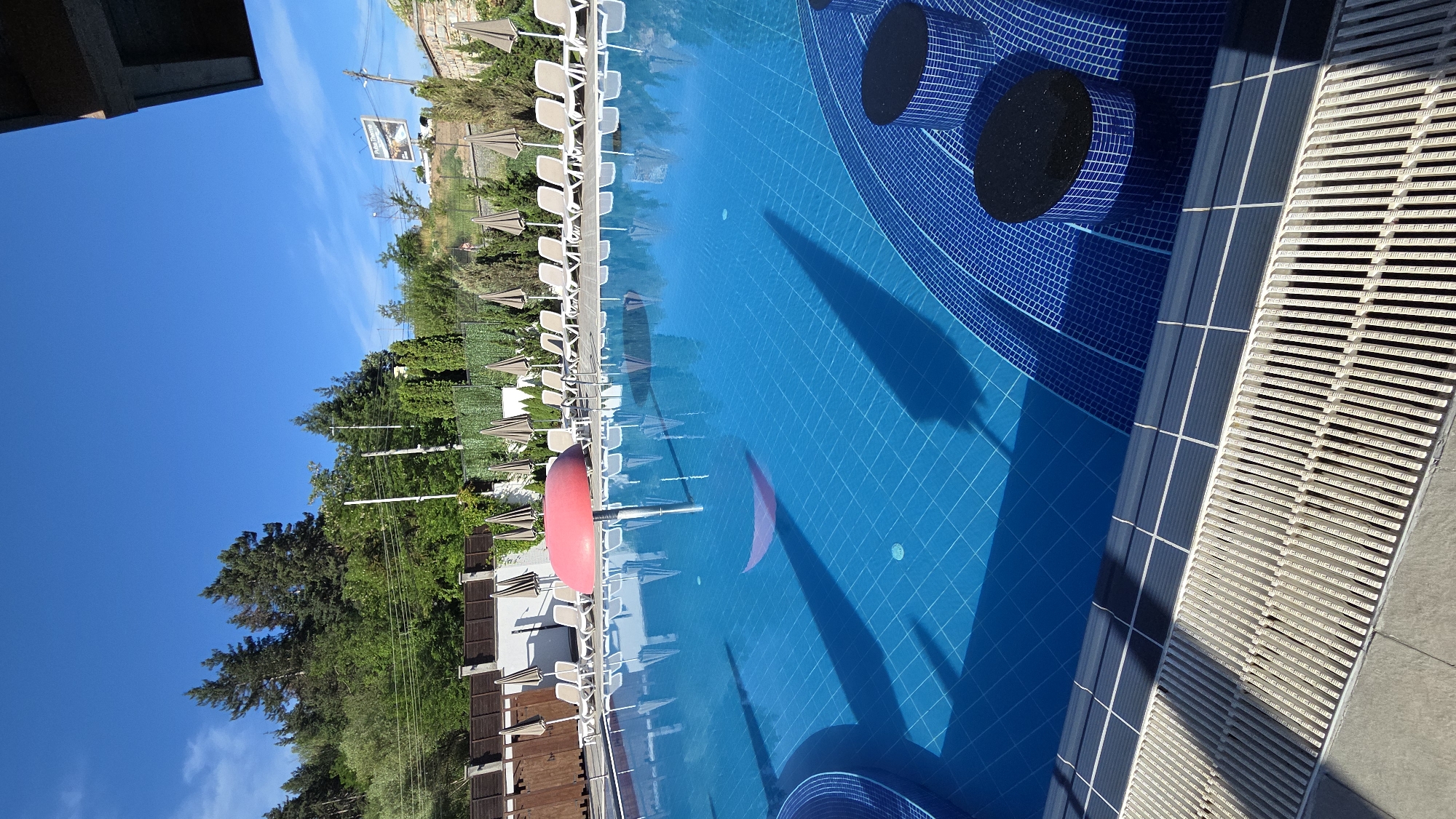 An outdoor swimming pool is pictured on a clear, sunny day. The pool's surface is bright blue and tiled, with a distinct checkered pattern visible. A large, red, mushroom-shaped structure is partially submerged in the water. Around the pool, rows of empty lounge chairs and closed umbrellas are arranged on a tiled deck. The deck is bordered by a white tiled edge and a drainage grate. In the background, a lush green hillside is covered with trees, and a fence and some buildings are visible. The sky above is a vibrant blue with a few wispy white clouds. The scene appears to be a tranquil resort or public pool area.