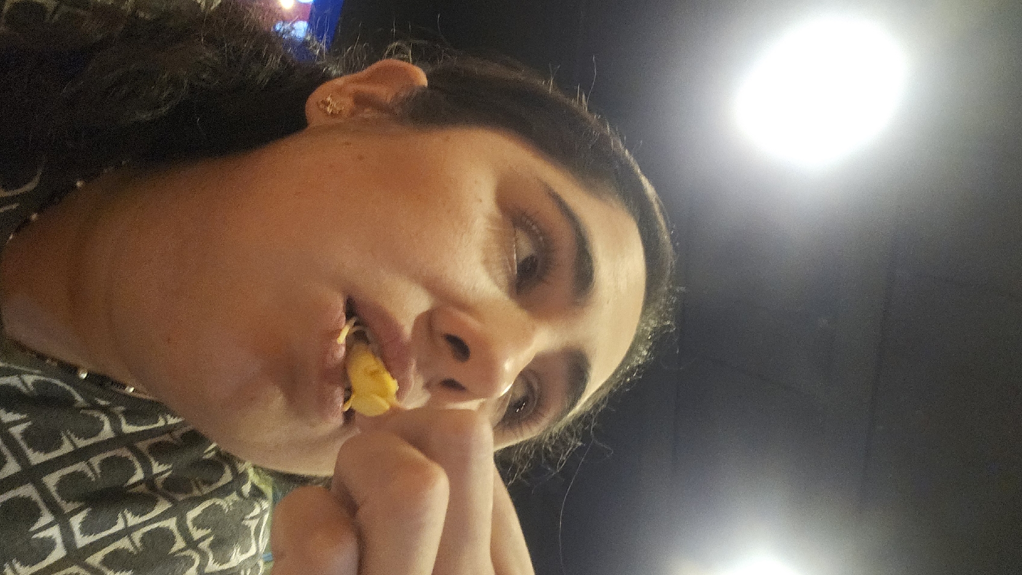 A medium, indoor shot depicts a woman, possibly in her late 20s, eating a yellow food item. Her face is turned slightly down and to the left, with her right hand near her chin. She has dark hair, an earring in her left ear, and is wearing a patterned shirt. The background is dark, with two lights: one appears brighter, indicating an overhead source, and the other less bright, possibly a light fixture. There is no indication of location in Puebla, Mexico.