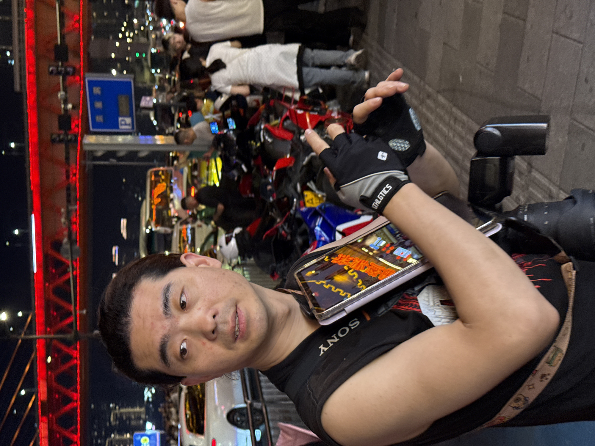 A selfie-style photograph shows an Asian man wearing a black Sony T-shirt with "ATHLETICS" on the sleeve and fingerless gloves, pointing towards a crowd of people lying on the ground. A smartphone resting on his shoulder displays a brightly colored image or game. The background includes a red metal structure, potentially a bridge or building frame, and several motorcycles or scooters. The scene is illuminated by streetlights and city lights, suggesting an urban environment at night. Based on the presence of the Chongqing University sign, the location is potentially in the city of Yuzhong District, China.