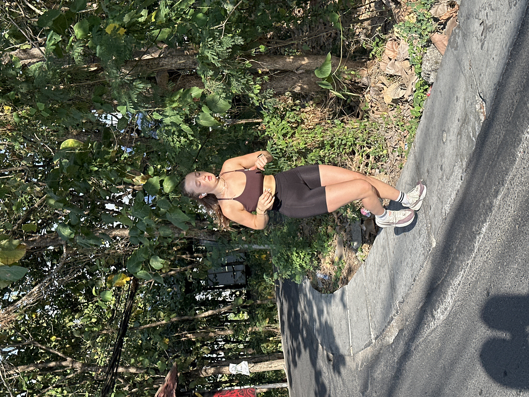 A young woman with blonde hair pulled back, wearing a dark brown sports bra and black shorts, is jogging on a paved path next to a road. She has white earbuds in and is wearing white athletic shoes with pink accents and white socks with black stripes. The scene takes place in North Kuta, Indonesia, characterized by lush green trees and vegetation lining the road and path. The sun is casting strong shadows on the road, indicating it is daytime. The road appears to be a street in a residential or semi-rural area, with a concrete curb separating it from the dirt and foliage. In the background, there are more trees and some electrical wires visible. The overall impression is of an outdoor exercise activity in a tropical environment.