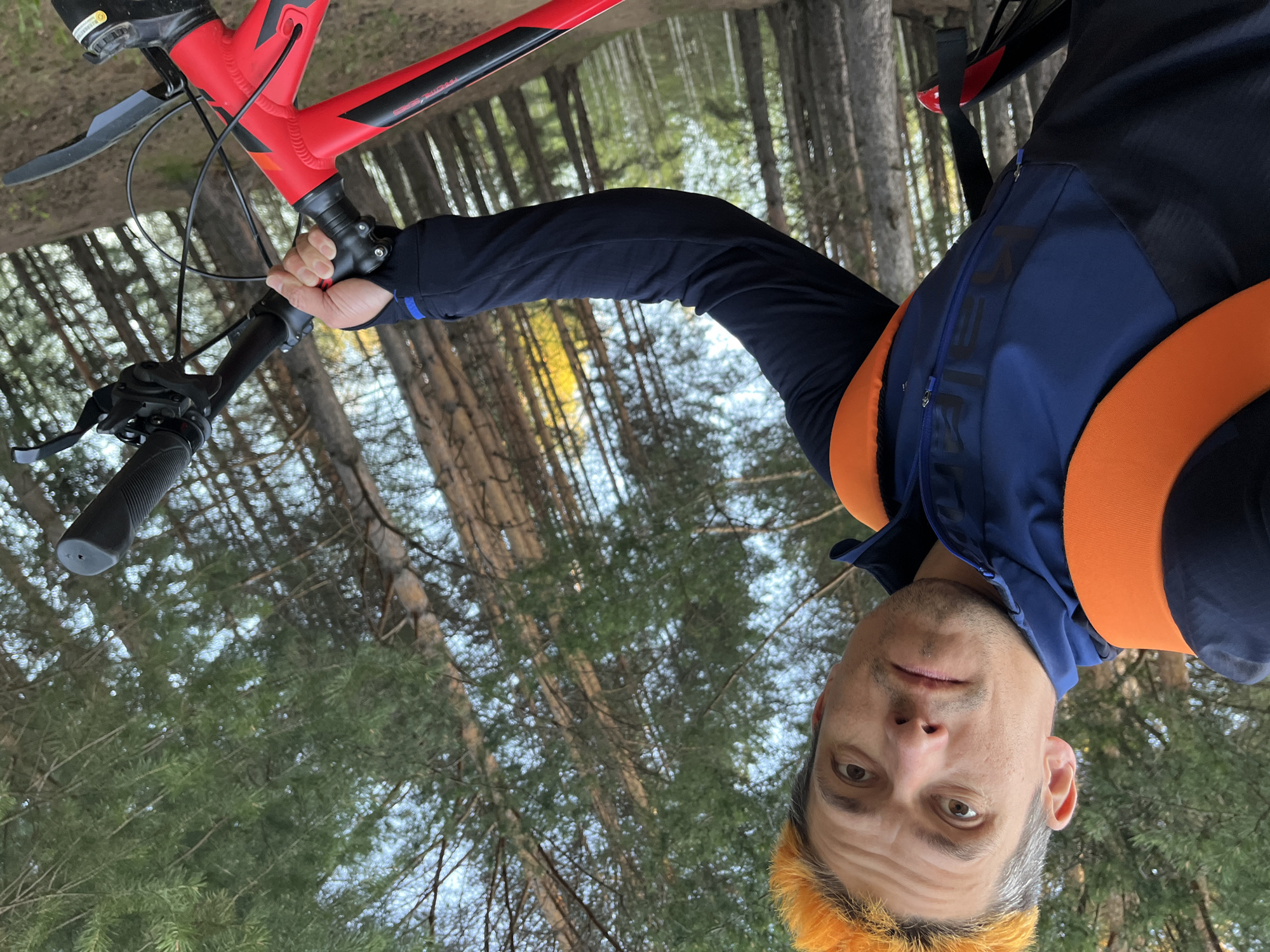 A man holds a red bicycle in a forest. The man is wearing a blue and orange jacket and has orange hair. The trees in the background are tall and green. The location is the city of Chaikovsky, Russia.