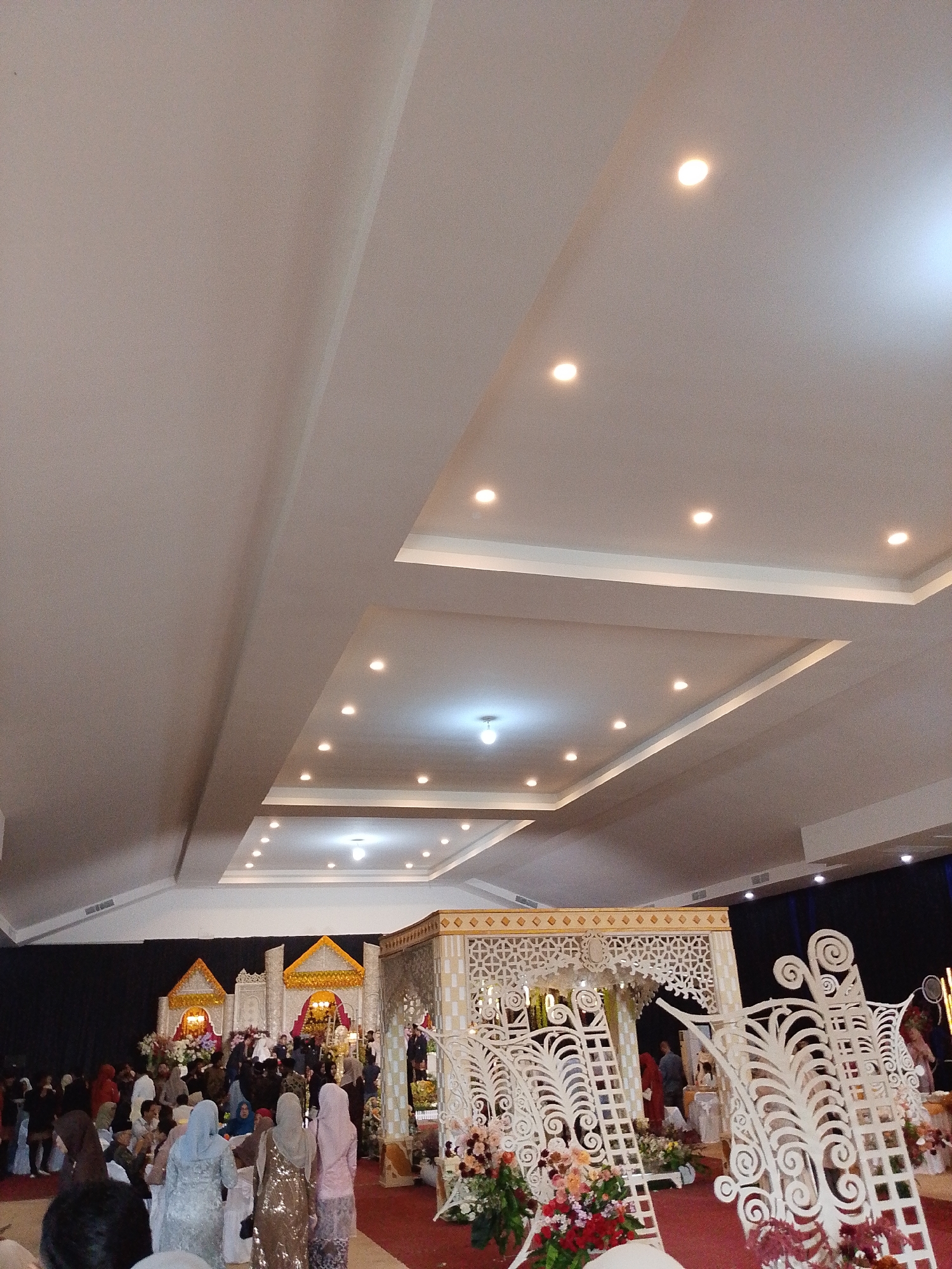This image depicts a wedding event in Lhokseumawe, Indonesia. Several people, many wearing traditional headscarves, are present in the room. The room is decorated with ornate, white-painted structures and floral arrangements. At the back, two golden and white canopies are visible against a backdrop of dark curtains. The ceiling is multi-tiered and features recessed lighting. A red carpet covers the floor.