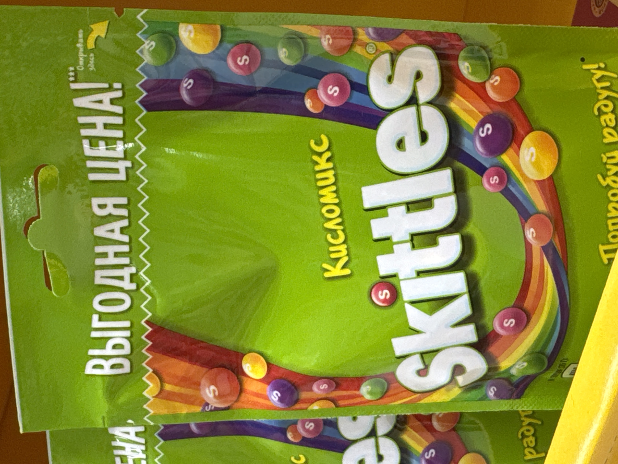 A close-up, slightly angled shot shows part of a package of Skittles candy. The packaging is a vibrant green, with the brand name "Skittles" prominently displayed in large, white, and slightly tilted letters. A rainbow arc of colors adorns the package, with individual Skittles candies integrated within the rainbow design. The text "Кисломикс" (Kislomiks) is printed in yellow next to the "Skittles" name. To the left, printed vertically in white, is the Russian text "ВЫГОДНАЯ ЦЕНА!" (VYGODNAYA TSENA!) which translates to "Good Price!". Text in Russian "Попробуй радугу!" (Poprobuj radugu!) translates to "Try the rainbow!". The top left of the package has a perforated edge suggesting where to open it.
The location "Ново-Никольское, Russia" cannot be confirmed based on the visual information provided.