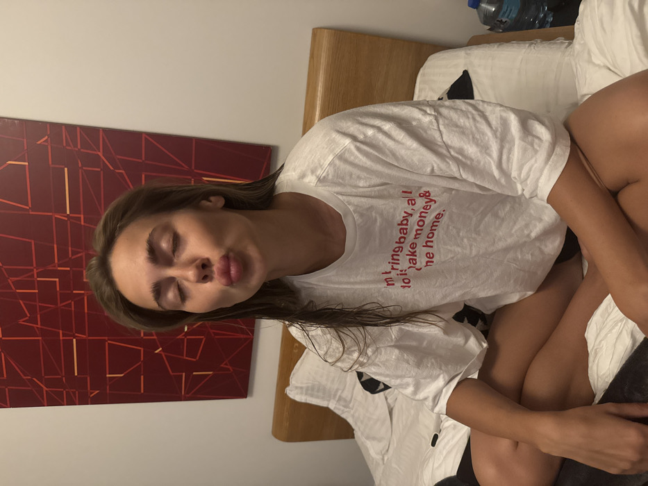 A woman in a white t-shirt with red lettering that reads "I don't bring baby, all I do is make money & come home" is lying in bed. She has light brown hair, pursed lips, and closed eyes. An abstract red geometric painting is behind her head. A water bottle is visible at the top of the bed. The room has neutral wall colors and light wooden furniture.