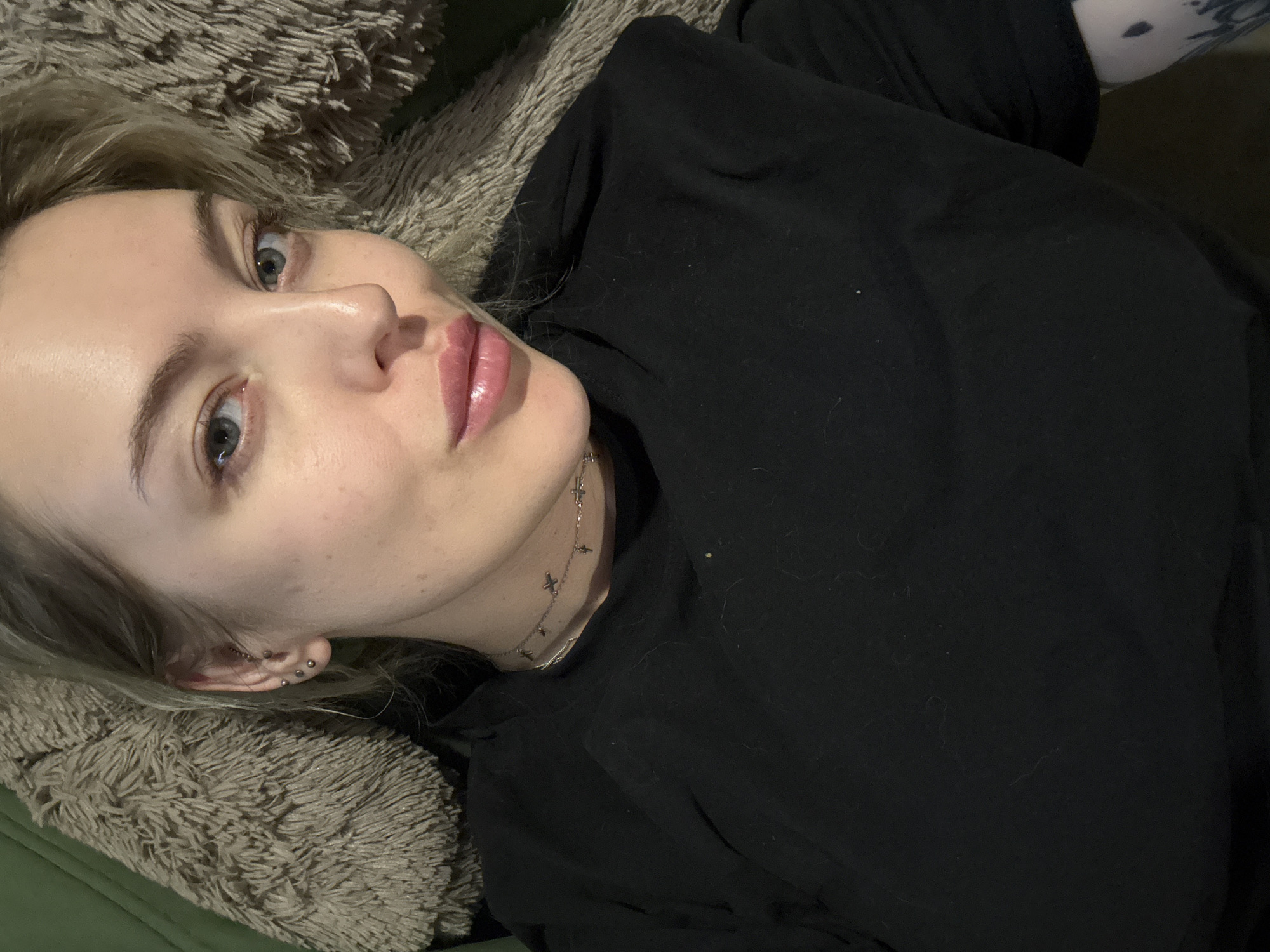 A fair-skinned woman with blonde hair is lying down on a light brown, textured cushion. She is wearing a black long-sleeved shirt and a silver necklace with cross pendants. She has multiple piercings on her left ear. Her gaze is directed towards the camera. Part of a green surface is visible beneath the cushion. The location of the image cannot be determined from the given information.