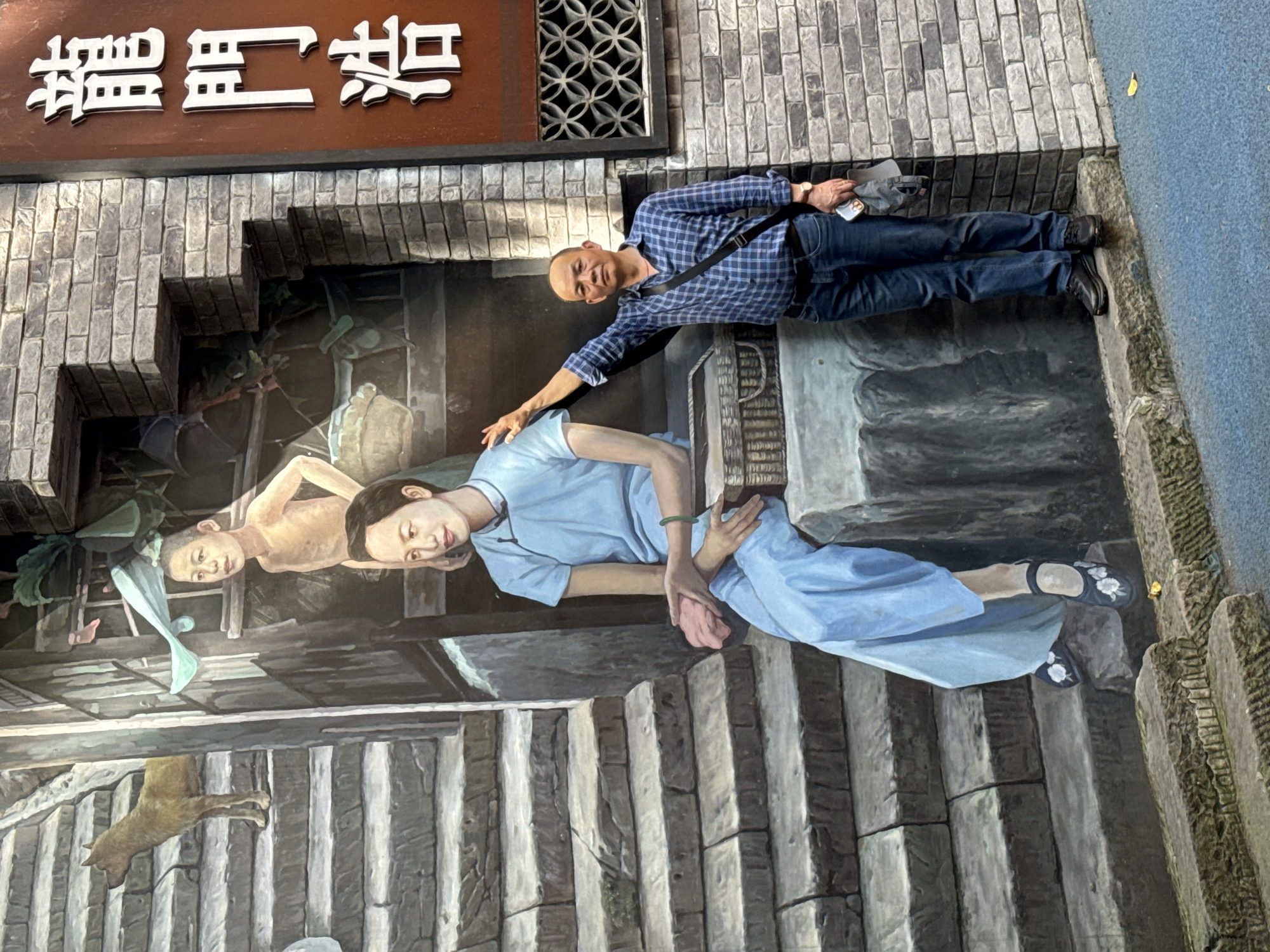 A vibrant outdoor scene captures a smiling bald man playfully interacting with a large 3D street art mural. The man, dressed in a blue plaid shirt, blue jeans, and dark shoes, stands on a blue paved surface alongside the artwork. He holds a smartphone in his right hand, wears a wristwatch and a black crossbody bag. With a joyful expression, he extends his left arm towards one of the painted figures, creating an engaging illusion of physical connection.
The elaborate mural, painted on a textured wall, depicts a realistic and detailed street or alleyway scene. In the foreground, a woman in a light blue dress with black embroidered shoes reclines on stone steps, looking upwards. She sports a green bracelet and holds a pink cloth. Behind her, a bare-chested young boy looks downwards, while another child's head is partially visible in the background. Towards the bottom left of the painted scene, an animal resembling a dog is shown on a wooden structure. The mural incorporates architectural details like brickwork, stone stairs, and an ornate grey patterned grate, enhancing its three-dimensional effect.
Above the mural, a prominent brown sign with raised white Chinese characters reads "貓兒莊" (Māo'er Zhuāng), which translates to "Cat Village" or "Cat Farm." The setting indicates a sunny daytime in an urban area. A small yellow leaf is visible on the blue pavement near the man's feet. This artwork, likely located in China's Nan'an District, transforms the street into an interactive visual experience, with the man's animated posture emphasizing the playful immersion it offers.