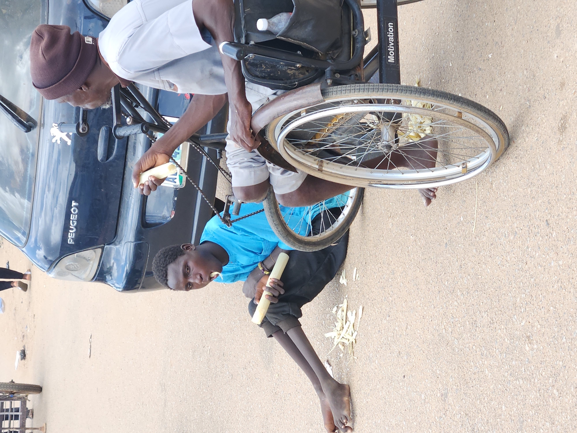 In Jos, Nigeria, a young person sits on the ground to the left of a wheelchair, chewing on a piece of sugarcane. The young person is wearing a bright blue tunic and black trousers, with bare feet. Pieces of chewed sugarcane are scattered on the ground around them. To the right, an adult, wearing a brown beanie and a light gray shirt, sits in the wheelchair, holding a piece of sugarcane and looking towards the left. The wheelchair has two large spoked wheels and a pocket on the side containing a plastic bottle. A dark blue Peugeot car is visible in the background, with its taillight and part of the trunk showing. The ground is paved with asphalt, typical of a street or roadside location in the city. The overall scene suggests a moment of rest or shared activity between two individuals in a public outdoor space in Jos.