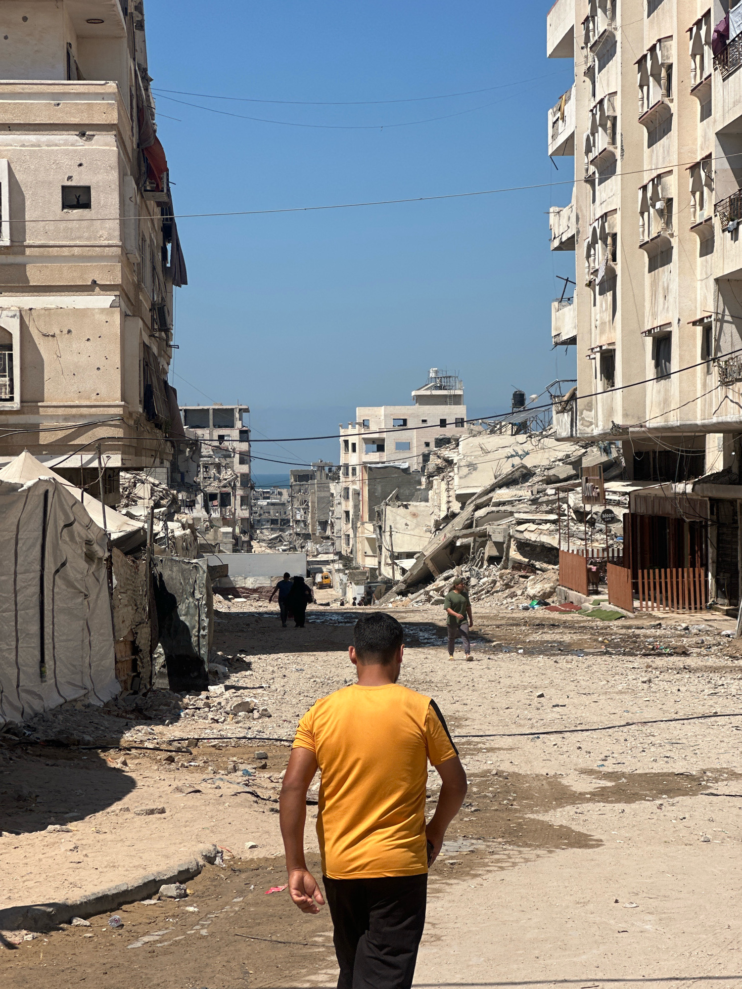 The image depicts a street scene in Gaza City, Palestinian Territories, characterized by widespread destruction. In the foreground, a person wearing a yellow t-shirt and black pants walks away from the camera. The street is unpaved and littered with debris, indicative of significant damage. On both sides of the street are multi-story buildings; those on the left appear to be partially intact with visible damage and bullet holes, while those on the right are severely damaged, with collapsed sections and rubble. In the distance, more damaged buildings and a clear blue sky are visible. Several other individuals can be seen walking in the midground and background, some near makeshift shelters or tents erected on the side of the road. The overall environment suggests a post-conflict or heavily damaged urban area.
