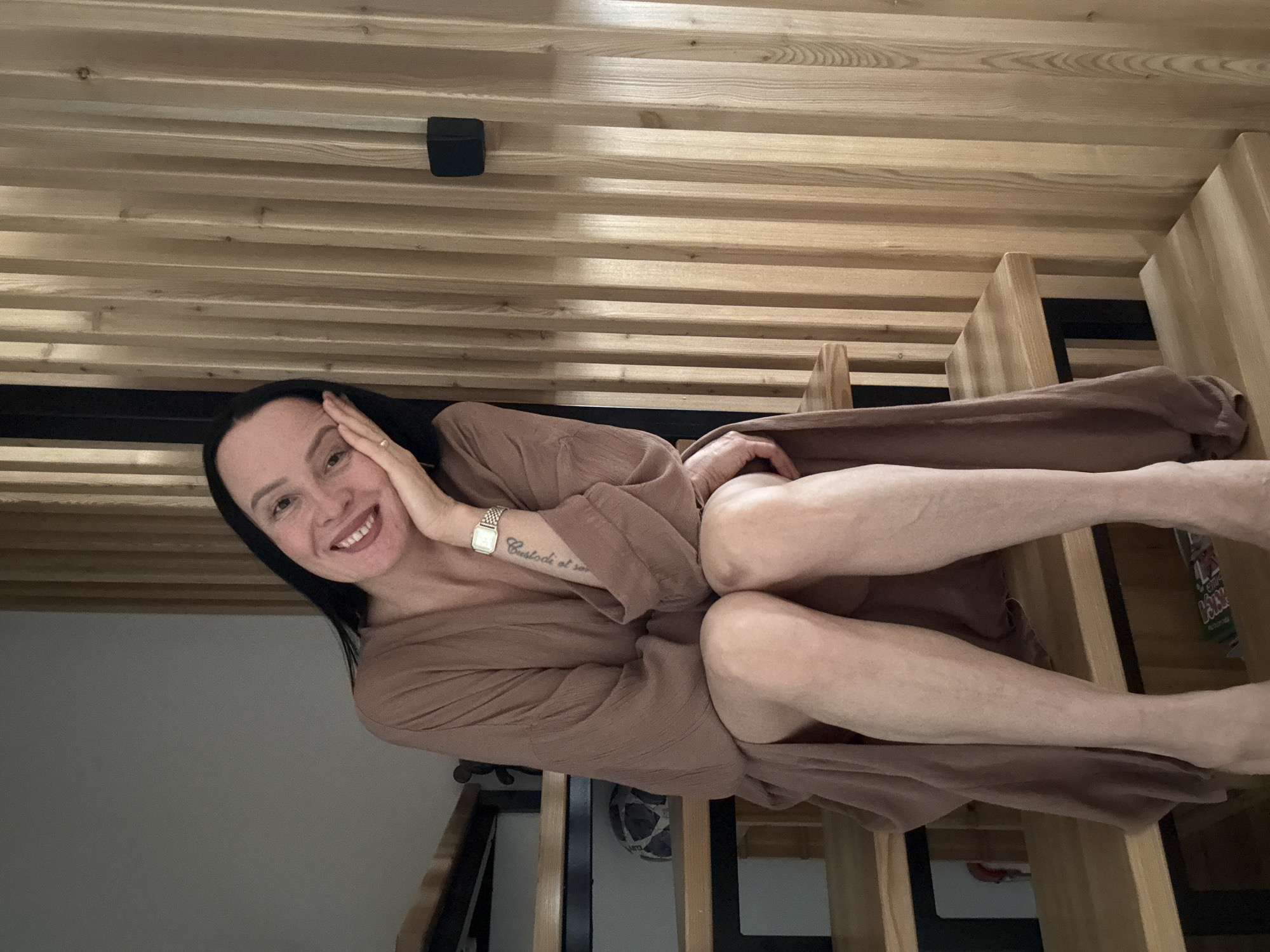A woman with black hair is smiling and reclining on a wooden staircase in a room with wooden paneling. She is wearing a brown robe and a watch on her left wrist. Her left hand is resting on her face. The location may be in Chaikovsky, Russia.
