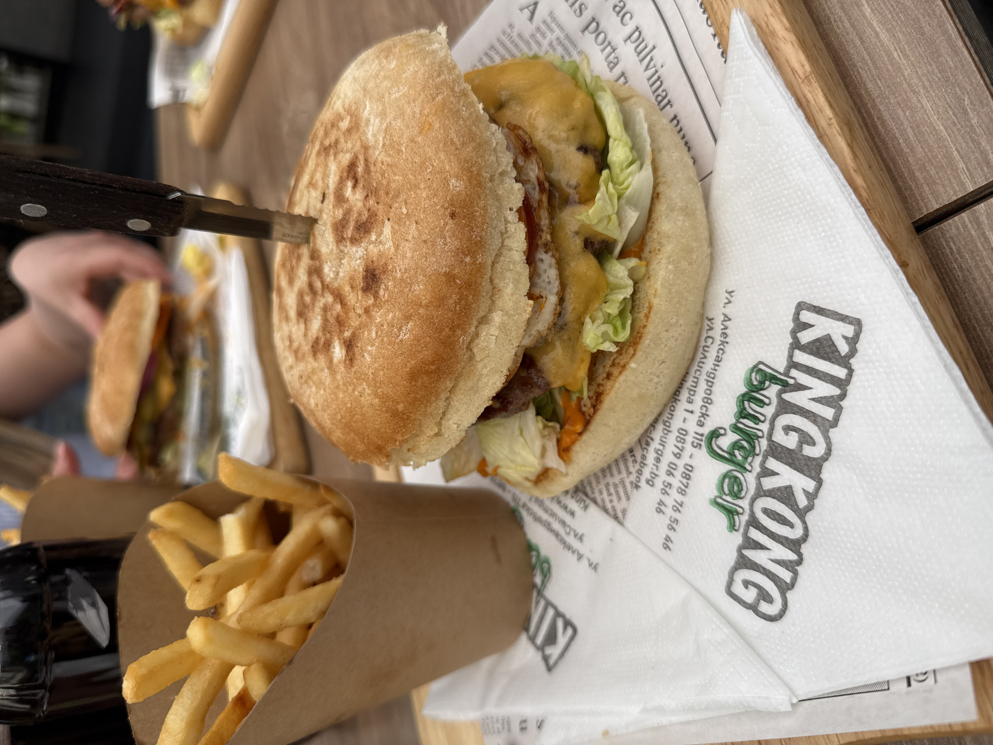 Here's a detailed description of the image:
The image shows a burger and fries on a wooden surface, likely a restaurant table. A knife is stuck into the burger. The burger appears to have a thick, round bun, and inside are visible layers of lettuce, cheese, and some kind of fried item, possibly an egg or chicken. Beside the burger is a paper cone filled with french fries. A napkin with the words "KING KONG Burger" is visible underneath the burger. The napkin also lists addresses on ул. Александровска and ул. Силистра in Burgas, Bulgaria, along with phone numbers and a website. In the blurred background, another burger is visible, and a person's hand appears to be reaching for it. The scene is well-lit, suggesting daytime.