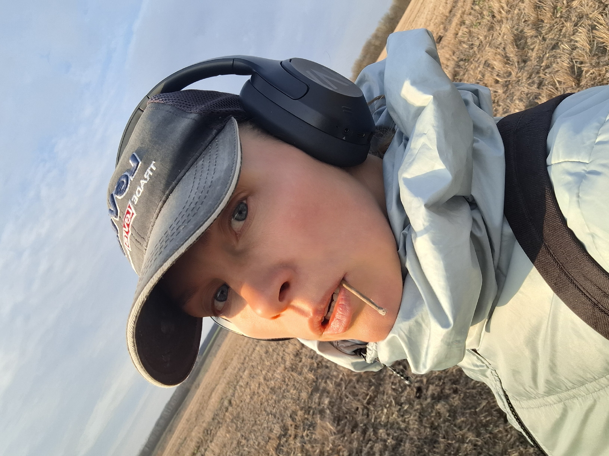 A person is centered in the frame wearing a gray baseball cap with red and blue lettering, black headphones, and a light blue hooded jacket. The person has a straw-like object in their mouth. The background shows a field of dried vegetation. The sky is partially visible with some clouds. Based on the description, this location is in the city of Khlevnoye, Russia.