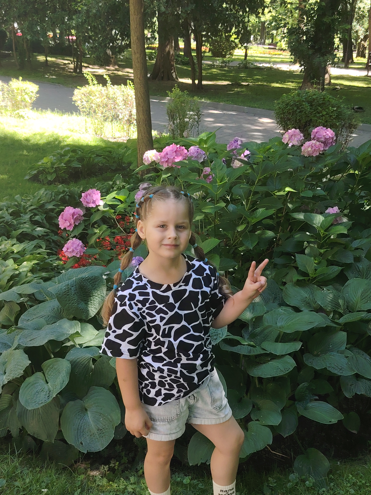 In the city of Homyel, Belarus, a young girl with braided hair stands in front of a lush garden. She is wearing a black and white patterned t-shirt and light denim shorts. Her right hand is raised, forming a peace sign. The garden is filled with large green hosta plants and clusters of pink hydrangeas, with some smaller red flowers interspersed. The background shows a tree-lined park with dappled sunlight on a paved path.