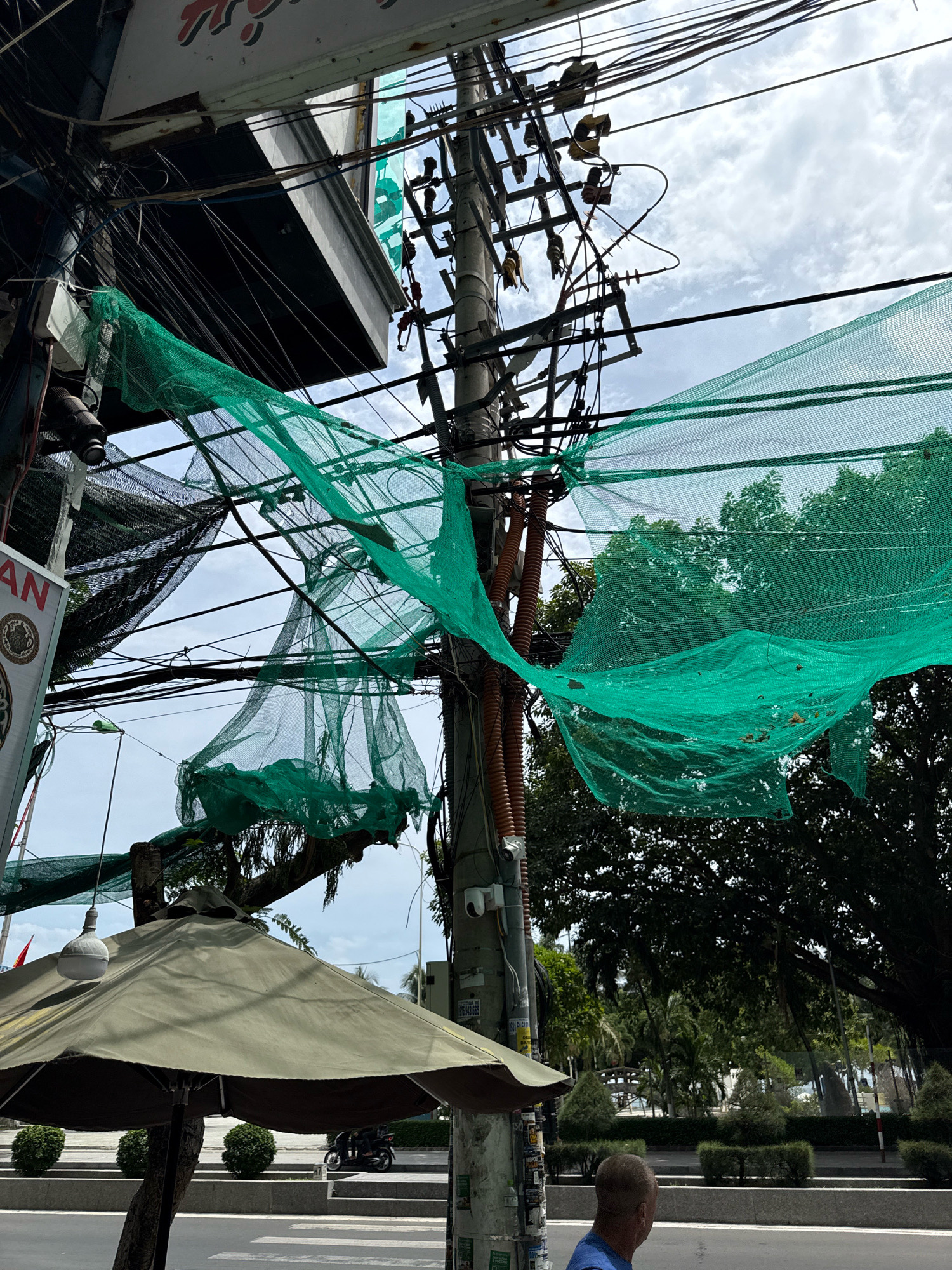 The image depicts a street scene in Nha Trang, Vietnam, characterized by a complex overhead network of electrical wires tangled with bright green safety netting. The netting, likely for construction or protection, is draped haphazardly over the wires, creating a chaotic visual. In the foreground, a large, olive-green umbrella provides shade, with a white light fixture hanging from its center. To the right of the umbrella, a utility pole with various attachments, including a security camera and numerous posters, stands prominently. Further down the street, a person in a blue shirt is visible from behind, facing away from the camera. In the background, lush green trees form a dense canopy, with hints of tropical vegetation. The sky is overcast with white clouds. The presence of electrical infrastructure, street furniture, and pedestrian elements suggests an urban environment typical of a coastal city.