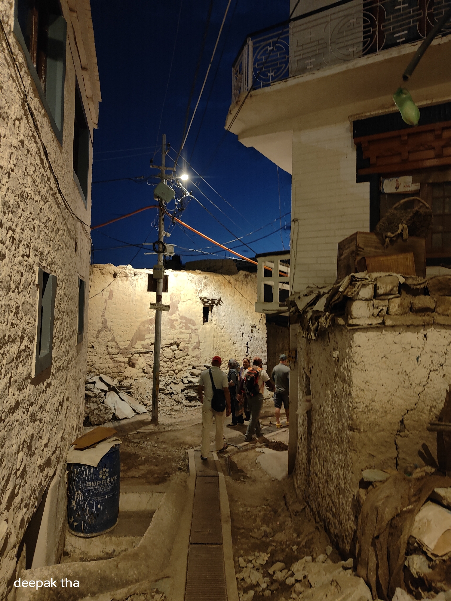 In the city of Leh, India, a narrow, dimly lit alleyway runs between two textured, light-colored buildings. The sky above is a deep twilight blue, with a cluster of overhead wires and a streetlamp illuminating the scene.
In the foreground, on the left, a large blue barrel with "SUPREME" printed on it rests in a recessed area. The ground is uneven, with dirt, rocks, and concrete elements forming the path.
Further down the alley, a group of five individuals are walking away from the camera. The person in front, a male, wears a red cap, a light-colored shirt, and khaki pants, carrying a backpack. To his left, a person in a gray t-shirt and shorts is also walking. Behind them, three more figures are visible, one wearing a backpack with an orange strap.
The architecture of the buildings suggests a traditional, possibly ancient, construction style, with rough plaster and visible stonework. A balcony with ornate metalwork is visible on the upper level of the building on the right. Debris and construction materials are scattered around, adding to the rustic atmosphere of the alley. The lighting creates long shadows and highlights the textures of the walls, contributing to a somewhat mysterious ambiance. The overall impression is of a quiet, residential area in Leh during the early evening hours.