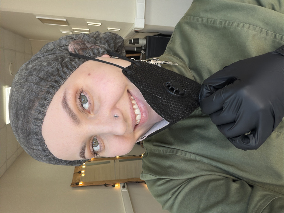 A person wearing a gray hairnet and black gloves smiles, holding a black respirator mask. A gun-shaped charm adorns the mask's strap. The person also has a silver nose piercing. They are wearing a green button-down shirt, presumably in a clinical or salon setting with a light wall and reflective surfaces in the background.