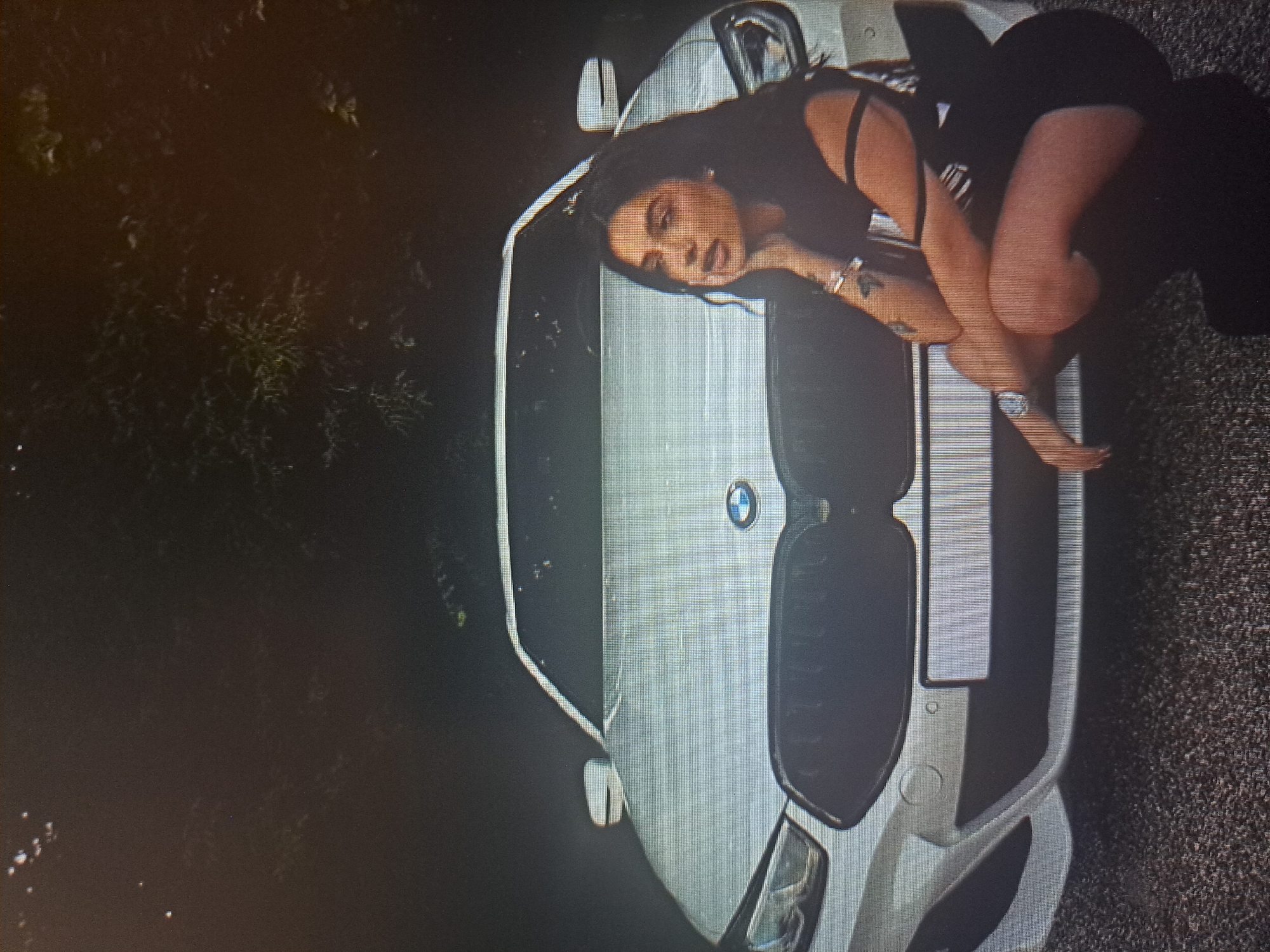 A woman is lying on the hood of a white BMW car. The car is parked on a dark paved surface, likely a road or driveway. The woman is wearing a dark outfit. The setting appears to be outdoors, with vegetation visible in the background. The exact location within Yerevan, Armenia, cannot be determined from the given image.