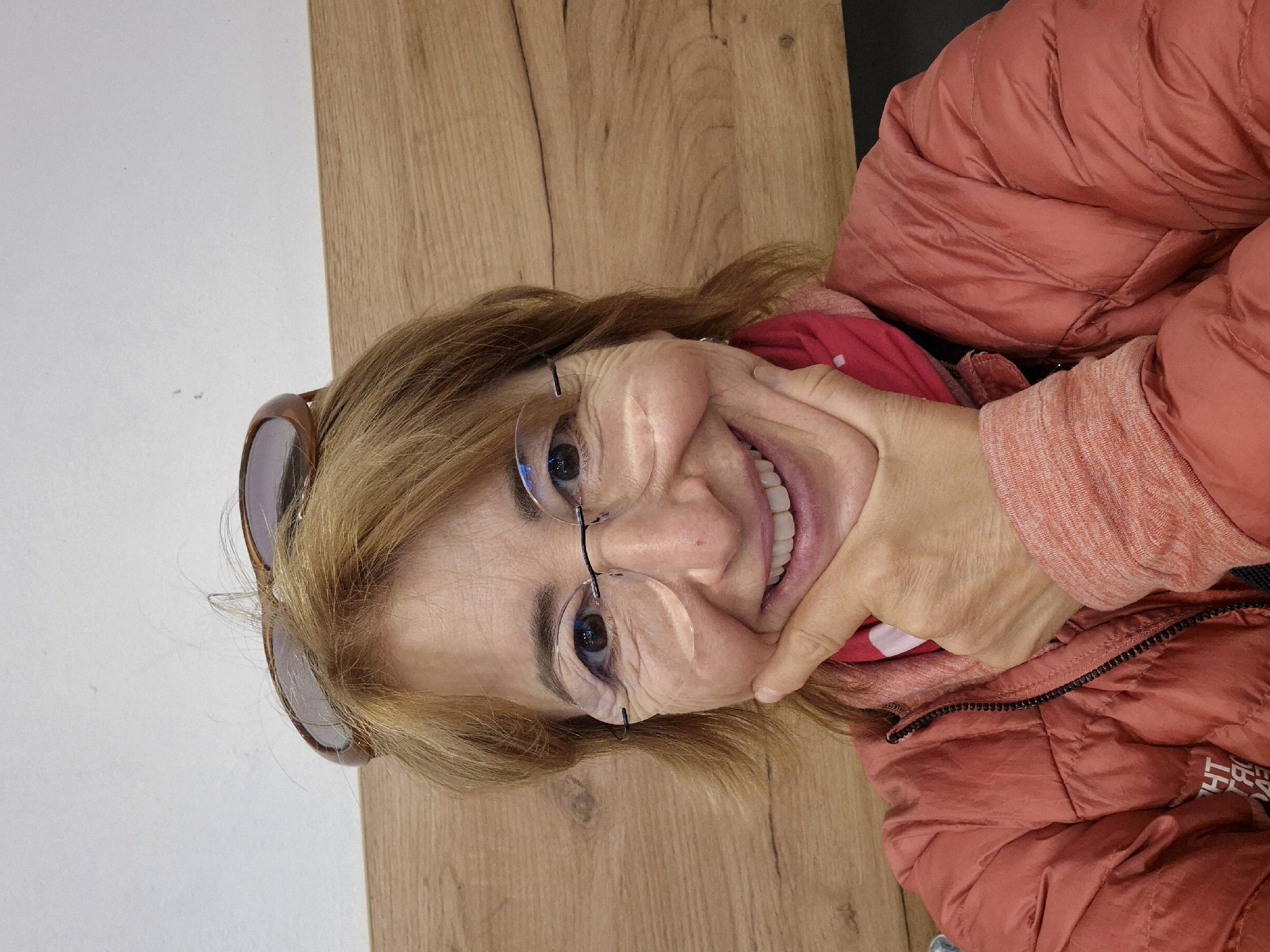 A close-up, portrait-style photograph shows an older woman with shoulder-length blonde hair smiling broadly. She is captured against a background split between a plain white wall on the left and a warm-toned wooden surface on the right, possibly a table or wall paneling, suggesting an indoor setting in Vienna, Austria.
The woman is wearing clear, round-lensed eyeglasses with thin dark frames, and a pair of brown-framed sunglasses are perched on top of her head. She is dressed in a reddish-coral puffy jacket, which has a black zipper partly visible on the right side of her chest. Underneath, a bright red garment is visible, and she wears a lighter, textured orange/coral long-sleeved shirt on her left arm. Her left hand is positioned on her chin and cheek, pressing lightly and creating folds in her skin, which highlights her wide, joyful smile, revealing her teeth. Visible wrinkles around her eyes and mouth convey her happy expression. Part of a logo, appearing to be "THE N F...", likely from "The North Face", is faintly visible on her jacket. The overall emotion conveyed is one of happiness and cheerfulness.
