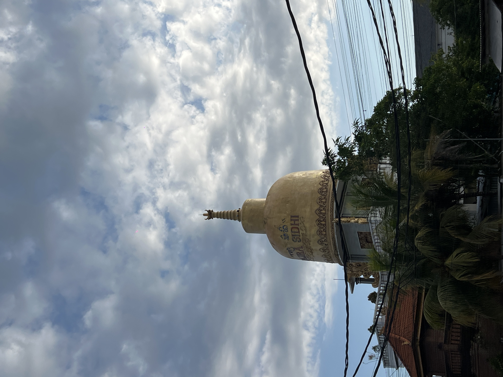 The image depicts a golden stupa, likely part of a Buddhist temple complex, in Mengwi, Indonesia. The stupa is partially obscured by trees and power lines. The sky is cloudy with patches of blue. Other buildings with red tile roofs are visible behind the trees. The stupa has an inscription that says "URA SIDHI".
