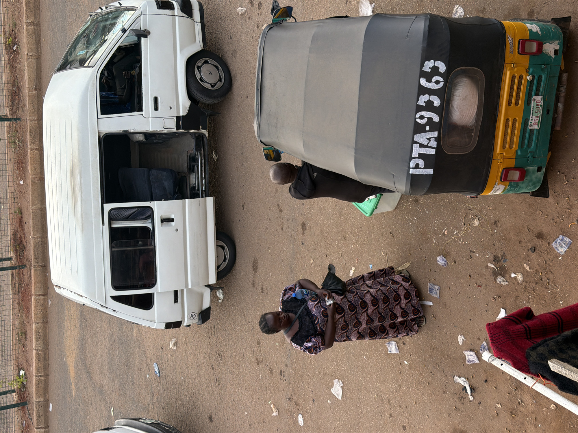 The image depicts an outdoor scene in Bukuru, Nigeria, featuring several vehicles and individuals on a dirt road. In the foreground, a three-wheeled auto-rickshaw, also known as a Keke Napep, with the license plate PTA-9363 visible, is parked. A man in a dark hooded sweatshirt is interacting with a green plastic container near the rear of the Keke Napep.
Behind the Keke Napep, a white minivan is parked. Its side door is open, revealing the interior with what appears to be dark seating. A woman in a long, patterned dress is standing to the left of the minivan, holding a plastic bottle and some currency. The ground is covered in dirt and scattered with pieces of litter. In the background, a metal fence and vegetation are visible, suggesting the edge of an urban or semi-urban area. The lighting indicates it is daytime.