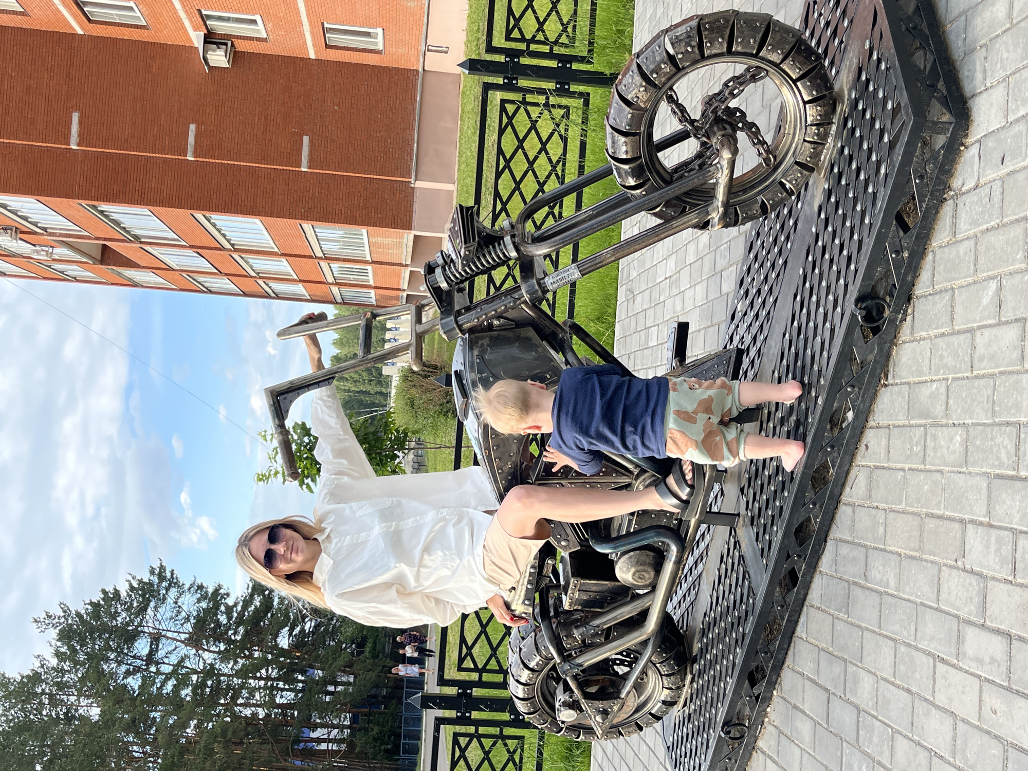 The image shows a woman sitting on a metal sculpture of a motorcycle, with a young child standing next to her. The woman is wearing sunglasses, a white oversized shirt, and light-colored shorts. The child is wearing a dark blue shirt and camouflage shorts. They are on a raised platform that is also made of metal, which sits on a paved area. In the background, there is a tall brick building with many windows, a black metal fence, and lush green trees. The sky is blue with some clouds. The setting appears to be an urban or semi-urban environment, possibly a public space or courtyard, located in the city of Chaikovsky, Russia.