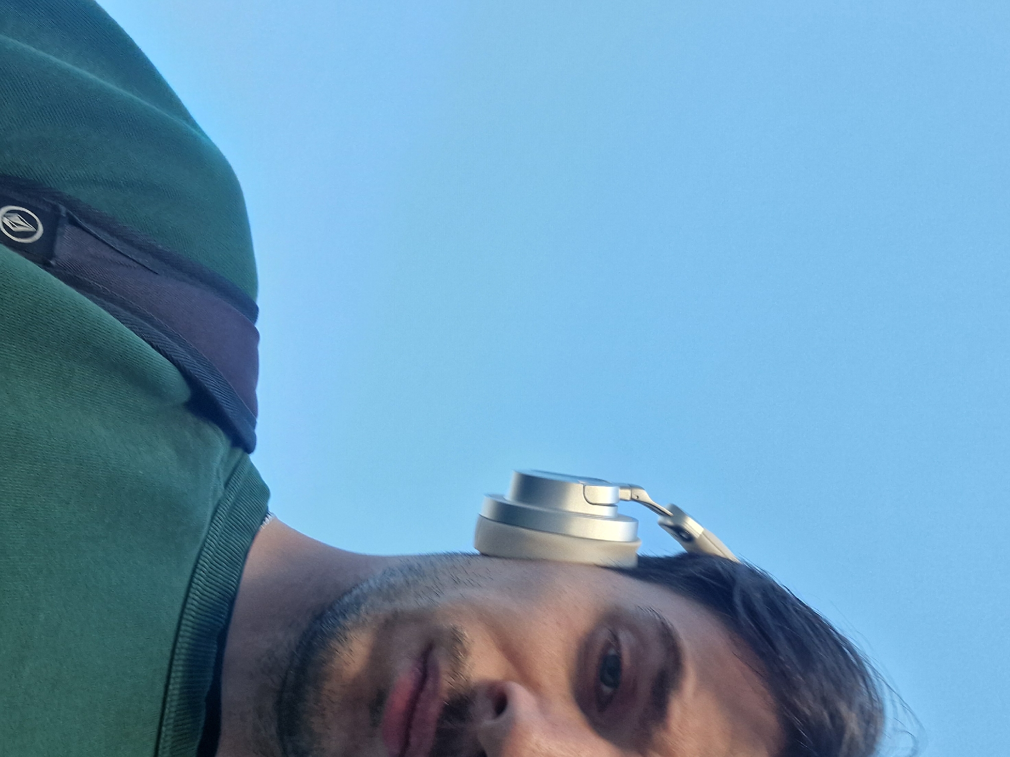 A low-angle, upward-looking photograph captures a person, likely male, against a bright and clear blue sky. The individual's head and upper body are positioned diagonally across the frame, with their face, featuring dark hair and stubble, oriented towards the bottom right. Their expression appears neutral or slightly relaxed.
A pair of silver or light grey over-ear headphones are resting on top of the person's head, rather than being worn over the ears. The person is dressed in a textured green garment, possibly a t-shirt or sweatshirt. A dark-colored strap or band, adorned with a circular logo, is visible across their shoulder or chest area, partially obscured by the green fabric. The logo features a distinct stylized diamond shape with a swirl, identifiable as the Volcom brand symbol. The uniform blue background indicates an outdoor setting during daytime with cloudless weather. The perspective suggests the photo may be a selfie taken from a low point of view.