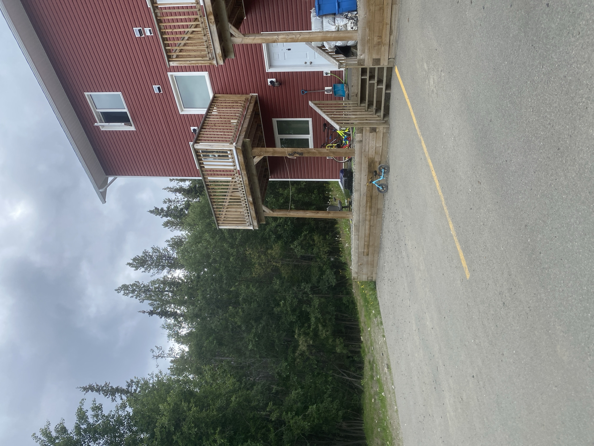 The image shows a red building with wooden balconies and stairs. A few windows are visible on the building's exterior. There are two bicycles near the stairs, one is blue and the other appears to be yellow and red. A blue bucket and a shovel are also present by the entrance. The building is situated next to a line of dense green trees. To the right of the building is a paved area with a yellow line marking. The sky is overcast, suggesting cloudy weather. The scene appears to be outdoors, possibly in a residential or recreational area in Whitehorse, Canada, given the context. There is no discernible activity or event taking place, and no text is visible in the image.