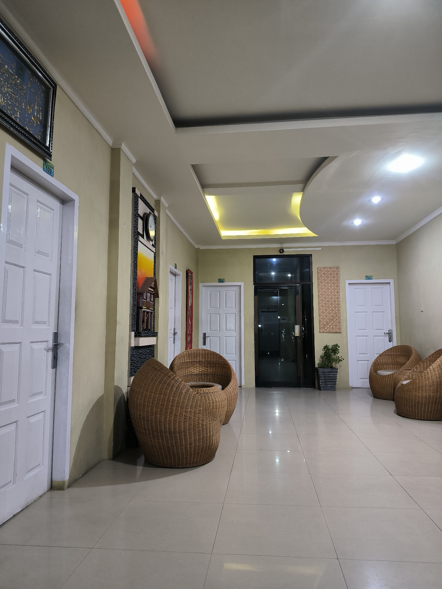 This media file captures an indoor hallway or waiting area, likely within a hotel, guesthouse, or apartment building in Banda Aceh, Indonesia. The space is well-lit by various artificial light sources, suggesting it is evening or night.
The main subjects include several white panel doors, three of which are clearly numbered "102," "104," and "105" with blue signs. A dark-framed glass door is positioned centrally on the right side of the image, possibly leading to an outdoor area or another section of the building. The walls are painted a light, warm beige or yellow color, complemented by white ceiling moldings and a multi-layered false ceiling featuring recessed spotlights and hidden strip lighting, some emitting a warm yellow glow, and a distinct red light accent on the left ceiling edge.
The floor consists of light-colored, glossy tiles, reflecting the overhead lights. A lounge area is furnished with several round, woven wicker armchairs, some appearing to have small light-colored cushions, arranged along both sides of the hallway.
Decorative elements include a framed piece of Arabic calligraphy on the left wall near door 102, a tall wall decoration featuring a clock and a miniature traditional house between doors 102 and the glass door, and a woven textured panel on the wall between the glass door and door 105. A small potted plant sits near the glass door, and a vertical red artwork is visible on the wall to the right of door 104. There are no people present, and no specific activity is taking place; the scene depicts a static, welcoming common area.