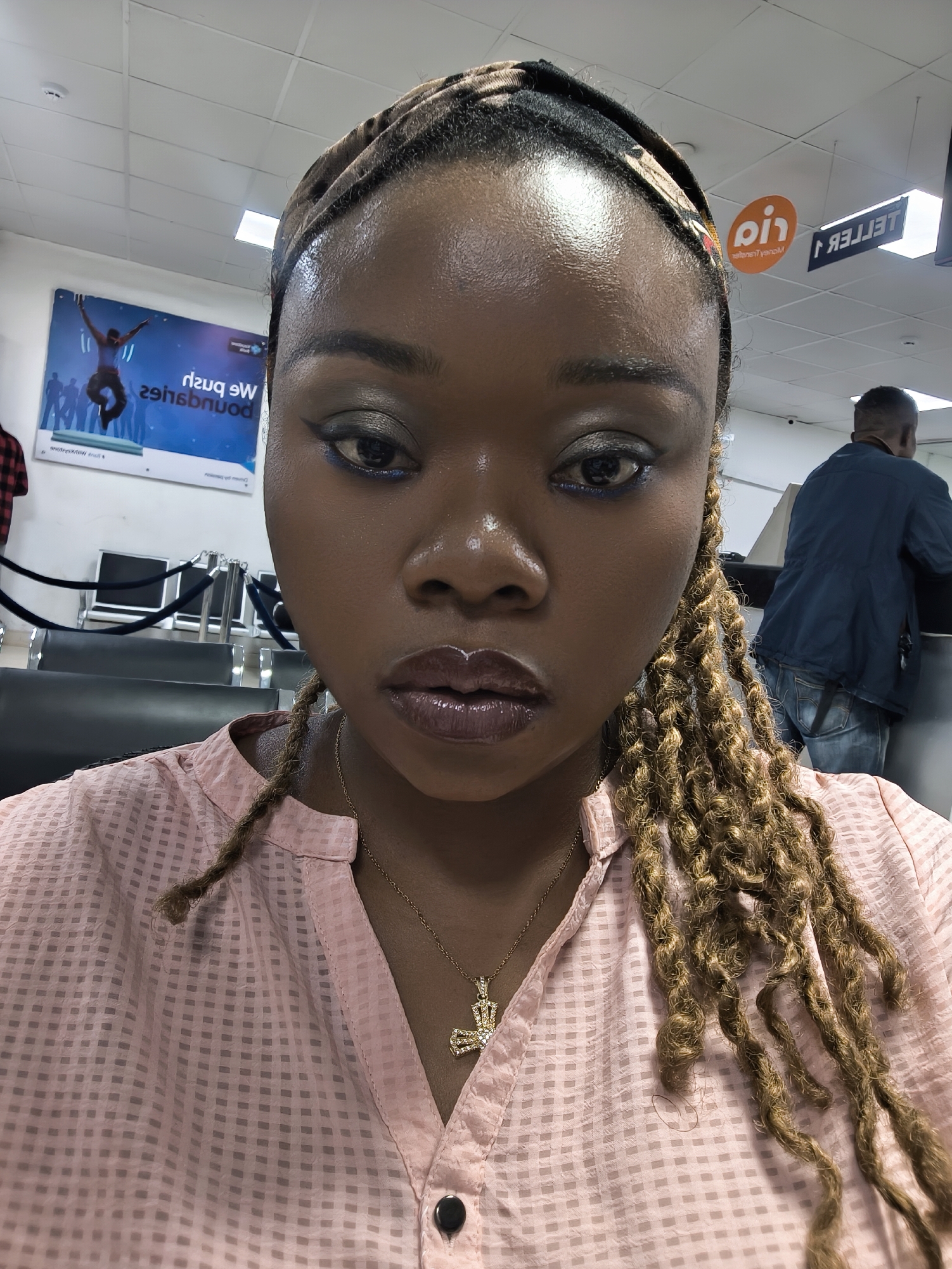 A medium close-up selfie photograph shows a woman with dark skin, wearing a patterned headband and locs. She is wearing a pink, checkered, button-down shirt and a gold necklace with a cross pendant. Her makeup includes glossy, dark-colored lipstick and heavy eyeliner. The background is an interior space, likely a bank or commercial establishment, indicated by the presence of teller signs, office furniture, and a poster advertising the bank's services. In the background, a man is standing at a counter. The location of this photograph is inferred to be in Jos, Nigeria.
