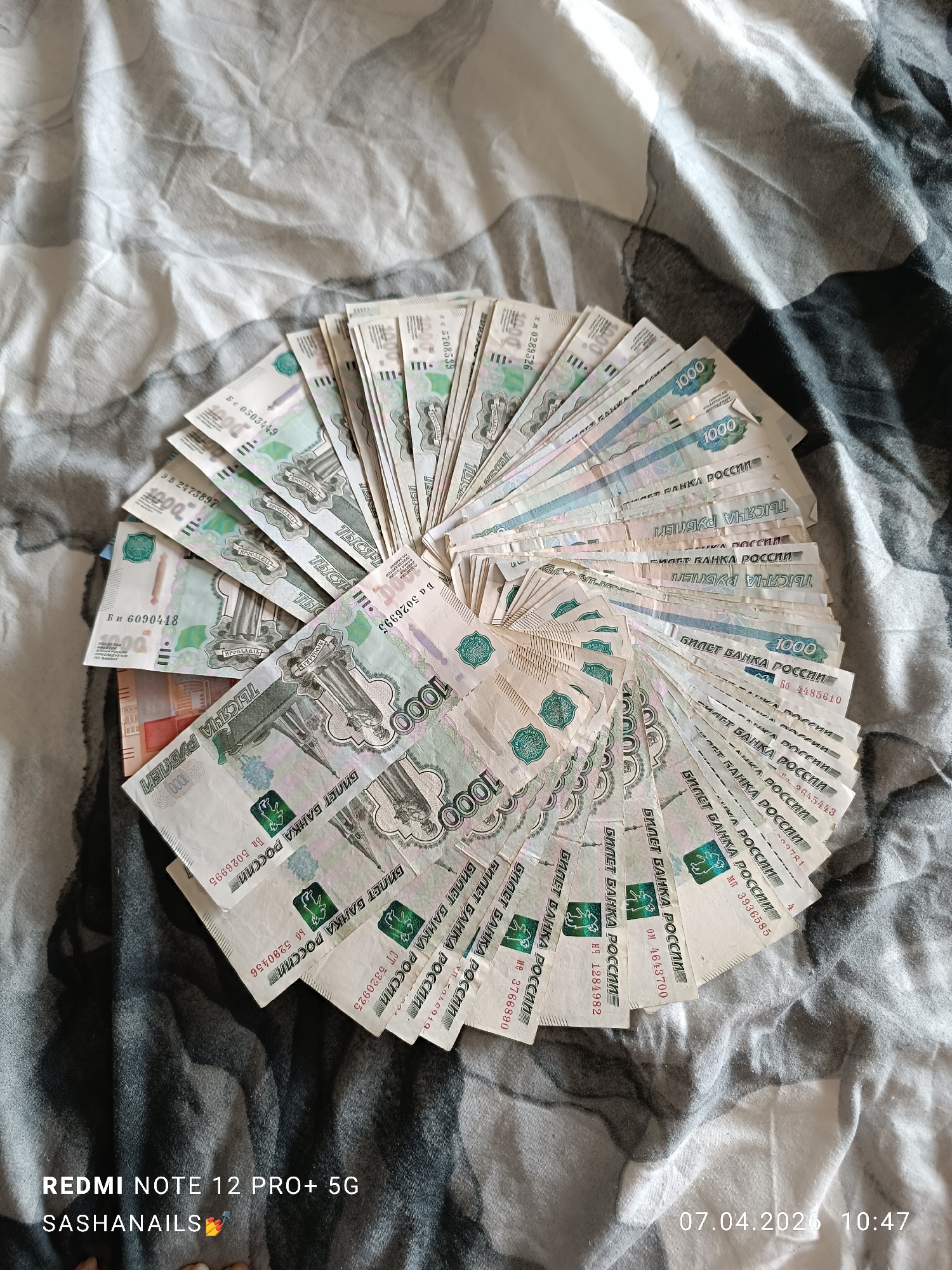 A high-angle shot shows a circular arrangement of Russian 1000 Ruble banknotes on a patterned cloth surface. The banknotes are predominantly white and green, and some are partially obscured. The cloth beneath has a predominantly gray and white pattern. In the lower left corner, there's a watermark indicating "REDMI NOTE 12 PRO+ 5G SASHANAILS." The lower right corner features a date and time stamp: "07.04.2026 10:47." There's no indication that the picture was taken in the city of Chaikovsky.