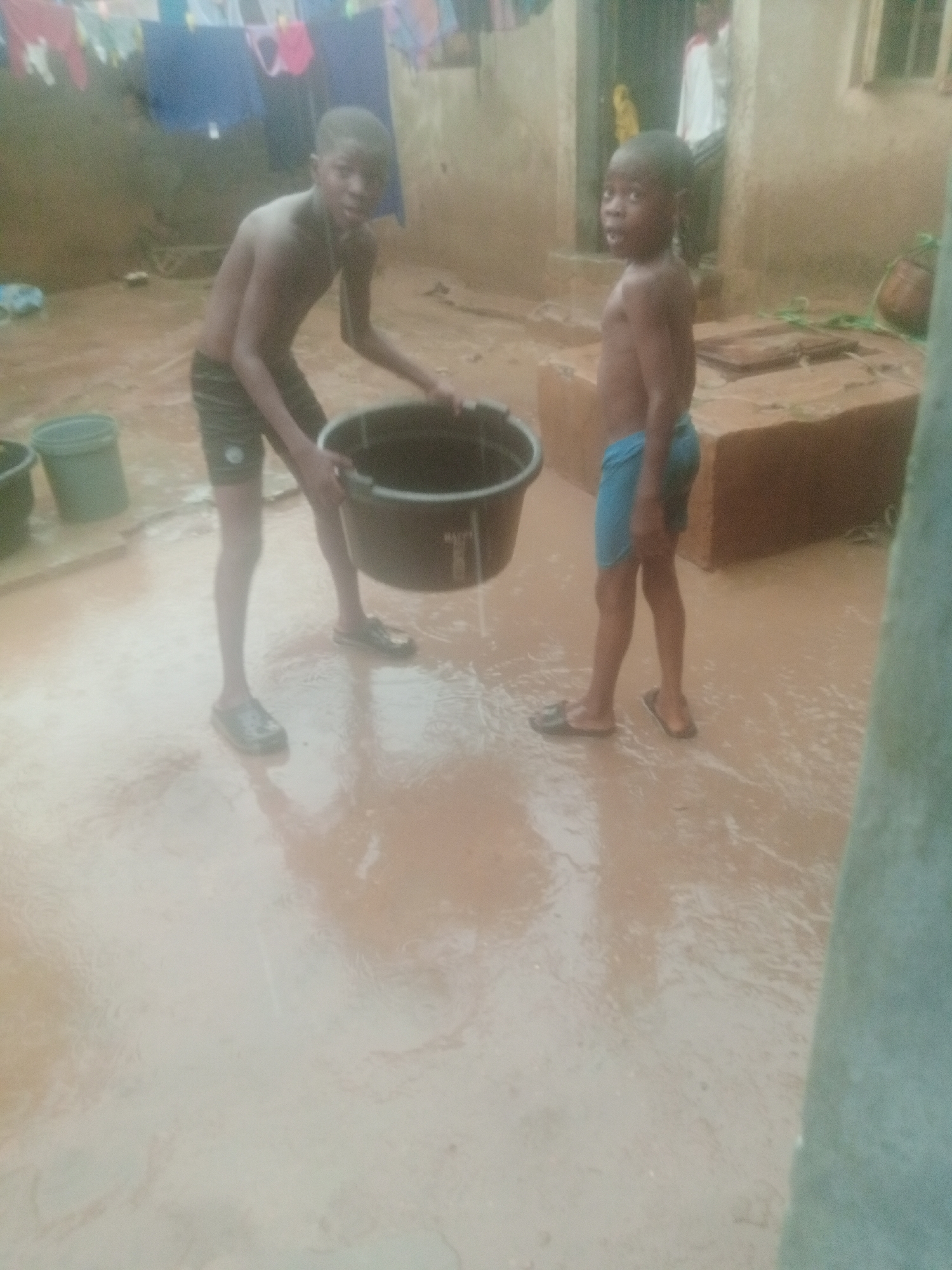 Two young boys stand in a muddy outdoor area. One boy, slightly older, wears dark shorts and black footwear. He is holding a large black bucket, seemingly full of water. The other boy wears blue shorts and sandals, standing to the side with a surprised expression. The ground is covered in a thin layer of reddish-brown water, creating a muddy appearance. In the background, there is a building with a weathered exterior and laundry hanging on a line. A rectangular brick structure is also visible behind the younger boy. The overall impression is of a casual scene in a rural or less developed environment. The image was taken in the city of Unknown, Unknown.