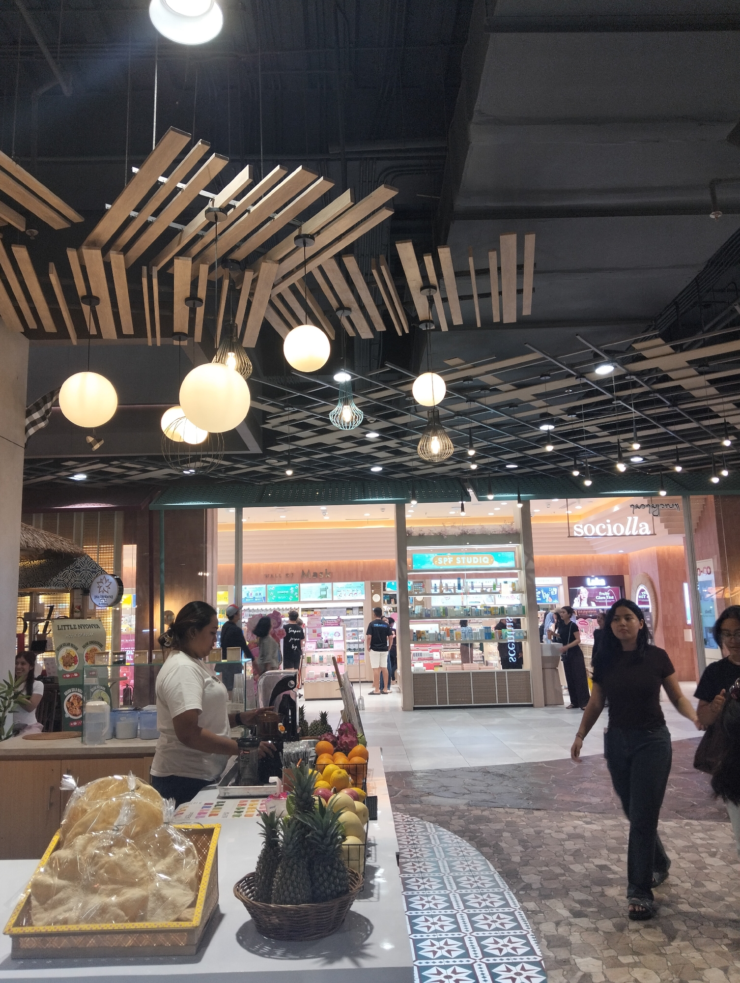 The image depicts the interior of a modern shopping mall, likely in Denpasar, Indonesia, given the signage. In the foreground, a stall displays various food items, including a basket of pastries or crackers wrapped in plastic, a woven basket with pineapples and other fruits, and a display of fruit. A woman in a white shirt is working behind the counter, operating a blender or juicer.
The background reveals a well-lit retail area with multiple storefronts. One prominent store has the word "sociolla" visible on its facade, and another displays "SPF STUDIO." Several people are seen browsing or walking through the mall, with some individuals in the foreground and others in the distance. The ceiling features an intricate design of wooden planks and various light fixtures, including large spherical lamps and smaller decorative ones. The flooring is a patterned tile, with a more ornate design near the foreground and a simpler pattern in the midground. The overall atmosphere is that of a busy commercial space.