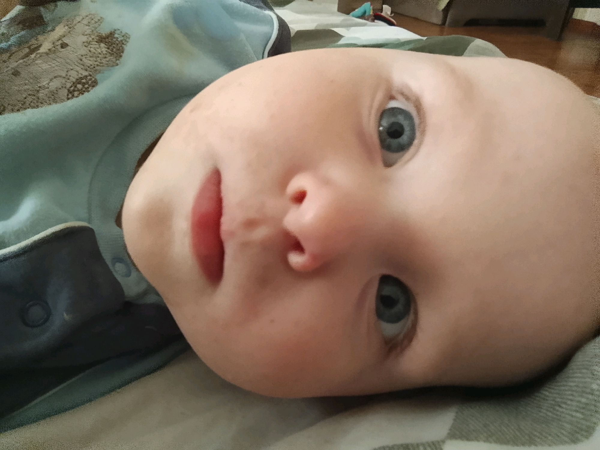 The image is a close-up, low-angle, eye-level shot of a baby's face. The baby, with fair skin and blue eyes, is lying on its back, facing towards the viewer. The baby is wearing a light blue garment with a patterned design on the left side. The background is a blurred surface, possibly a blanket or bedding, with a checkered pattern of white and grey. The lighting suggests an indoor setting. There are no discernible geographical landmarks that would indicate a location in Hrodna, Belarus, or any other specific city.