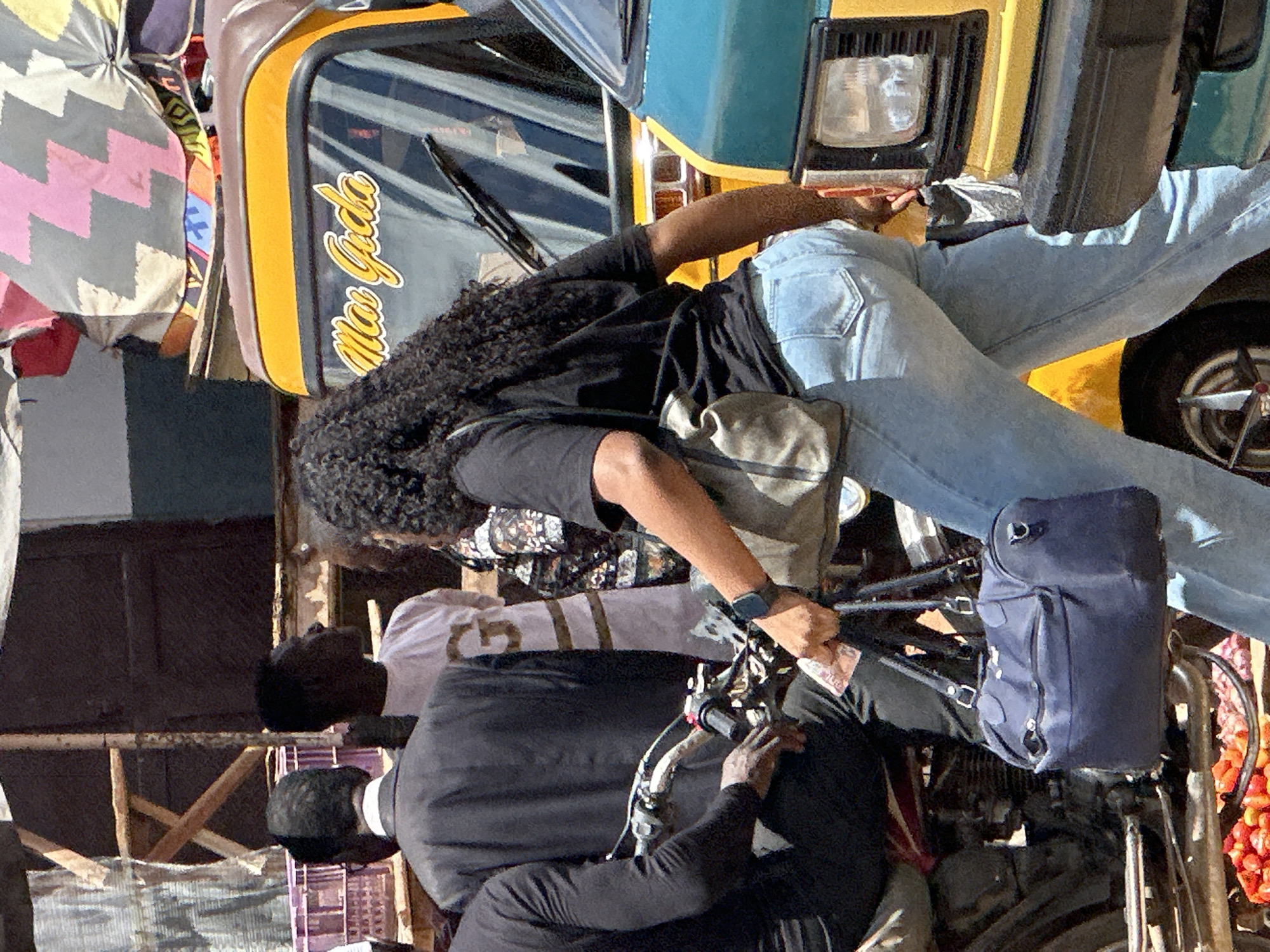 In Narayi, Nigeria, a street scene unfolds with a woman in light blue jeans and a black t-shirt holding a dark blue handbag and a light-colored purse. She stands near a yellow and brown auto-rickshaw with the inscription "Mas Gida" on its windshield and next to a teal vehicle. Behind her, a man in a black suit and another man in a light-colored garment are visible. The ground in the foreground displays a pile of red fruits, possibly tomatoes, and other produce. The background suggests an outdoor market or roadside setting with structures and umbrellas.
