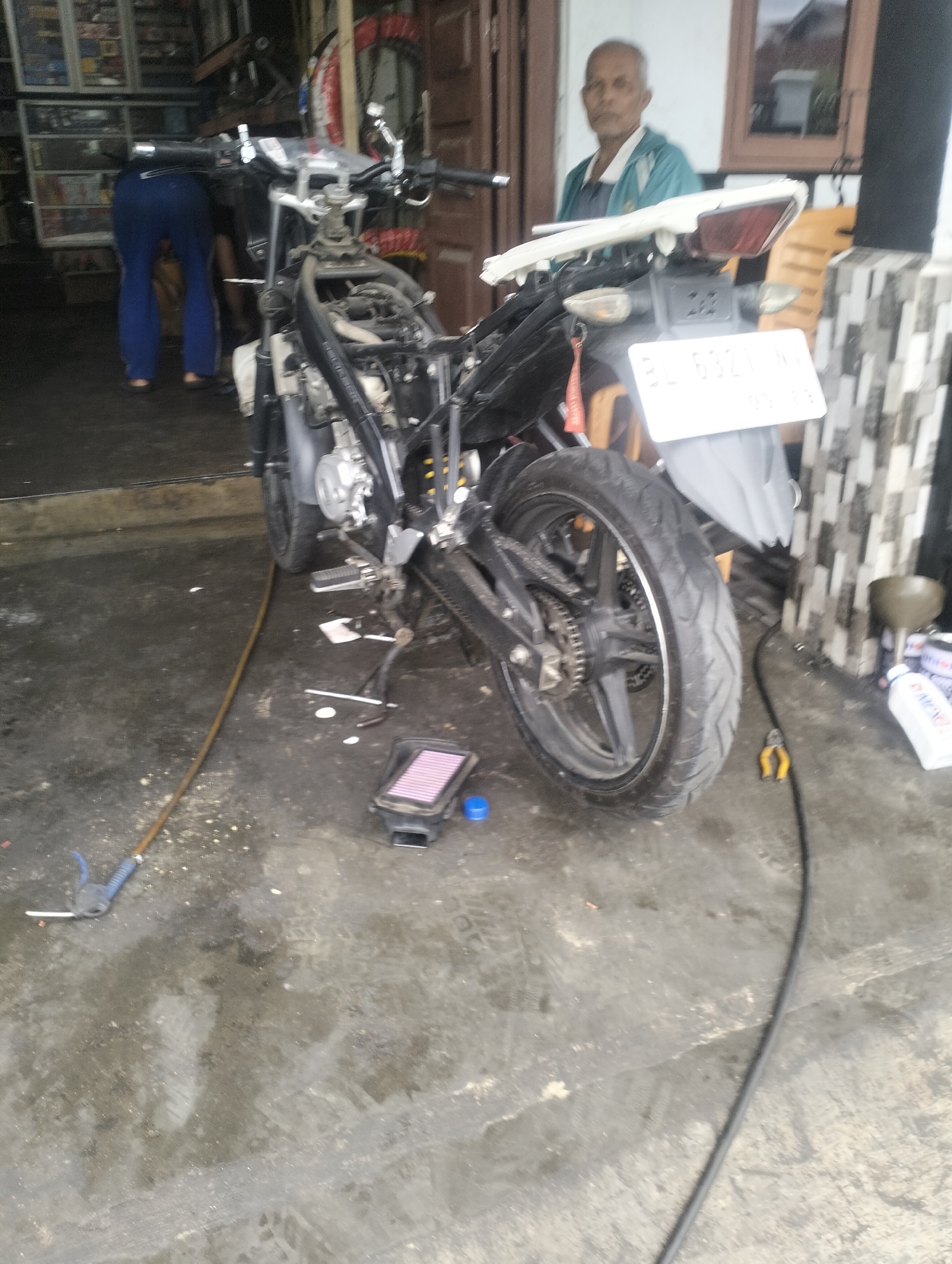 In Lhokseumawe, Indonesia, a motorcycle is being worked on in what appears to be a repair shop or a residential area with a concrete floor. The motorcycle, a sportbike, is partially disassembled, with its fairings removed. A man, likely the owner or mechanic, is standing near the rear of the motorcycle, observing. His face is obscured by shadows, but he is wearing a light-colored shirt and dark pants. In the background, shelves filled with various items, possibly motorcycle parts or other goods, are visible. A person in blue pants is bent over and appears to be working on something to the left of the motorcycle. The environment is cluttered with tools and debris, suggesting an active workspace.