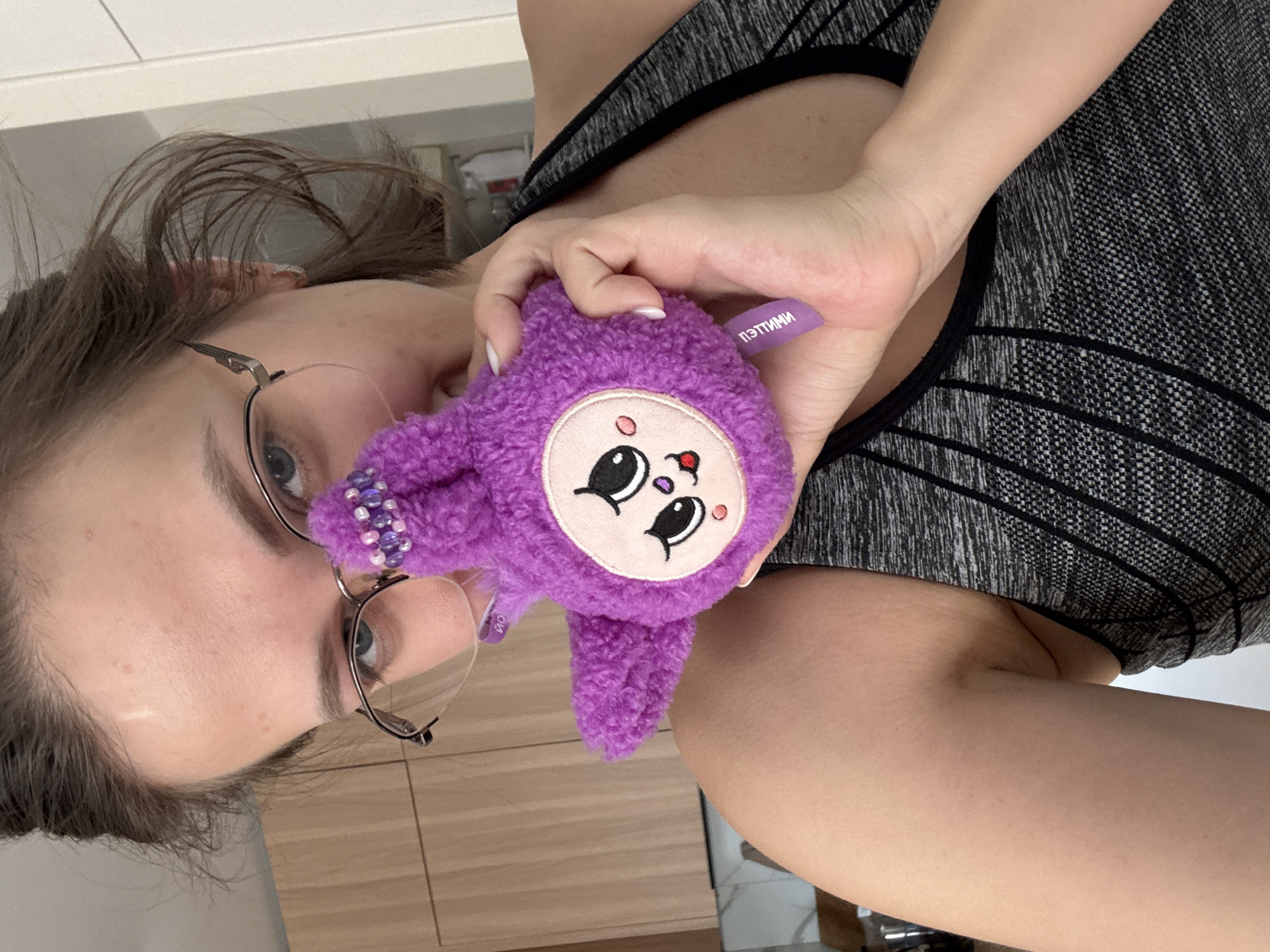 The image captures a close-up, sideways view of a woman indoors, holding a small purple plush toy. The woman is positioned on the left side of the frame, with her head, shoulders, and arm visible. She wears glasses with thin metal frames and has dark hair, some strands appearing loose around her face. Her expression is neutral, and her eyes are light-colored. She is wearing a grey and black patterned sports bra or top. Her arm, with fair skin, extends into the frame from the right, holding the plush toy in her hand, which has neatly manicured fingernails with light-colored polish.
The main object is a small, vibrant purple, fluffy plush toy with a circular light-colored face. The toy features large black eyes, small pink cheeks, a small pink nose, and a tiny red mouth or tongue. Near the top left of the toy's head, there's a small cluster of purple and clear beads, possibly attached to a loop. A purple fabric tag is visible on the toy, bearing the white text "ПЭТТИМИ" (PETTIMI).
The background is simple, suggesting an indoor setting with light-colored walls or cabinetry and a surface that appears to be light wood grain. The lighting is bright and even, indicating daytime. The overall mood is casual. The image contextually originates from Pervomaysky, Russia.