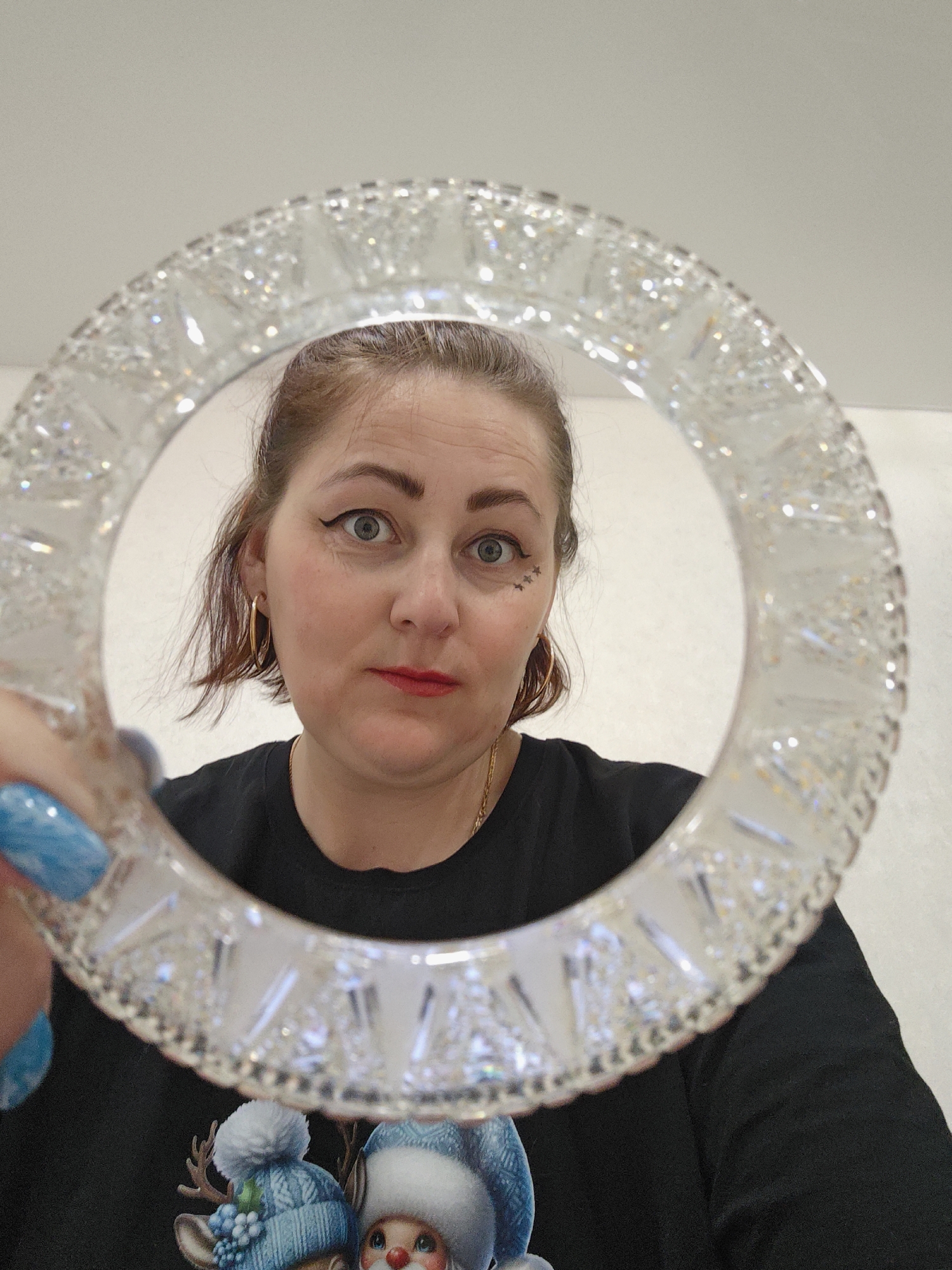 A woman with fair skin, brown hair, and bright red lipstick is looking directly at the camera through a decorative glass ring. She has eyeliner and small star-shaped facial tattoos near her left eye. She wears gold hoop earrings and a black t-shirt with a printed design of what appears to be winter characters and decorations. Her nails are painted light blue.
The background is a plain, light-colored wall. The location is likely indoors. There is no definitive information in the image to indicate the specific location is Markovskii, Russia.