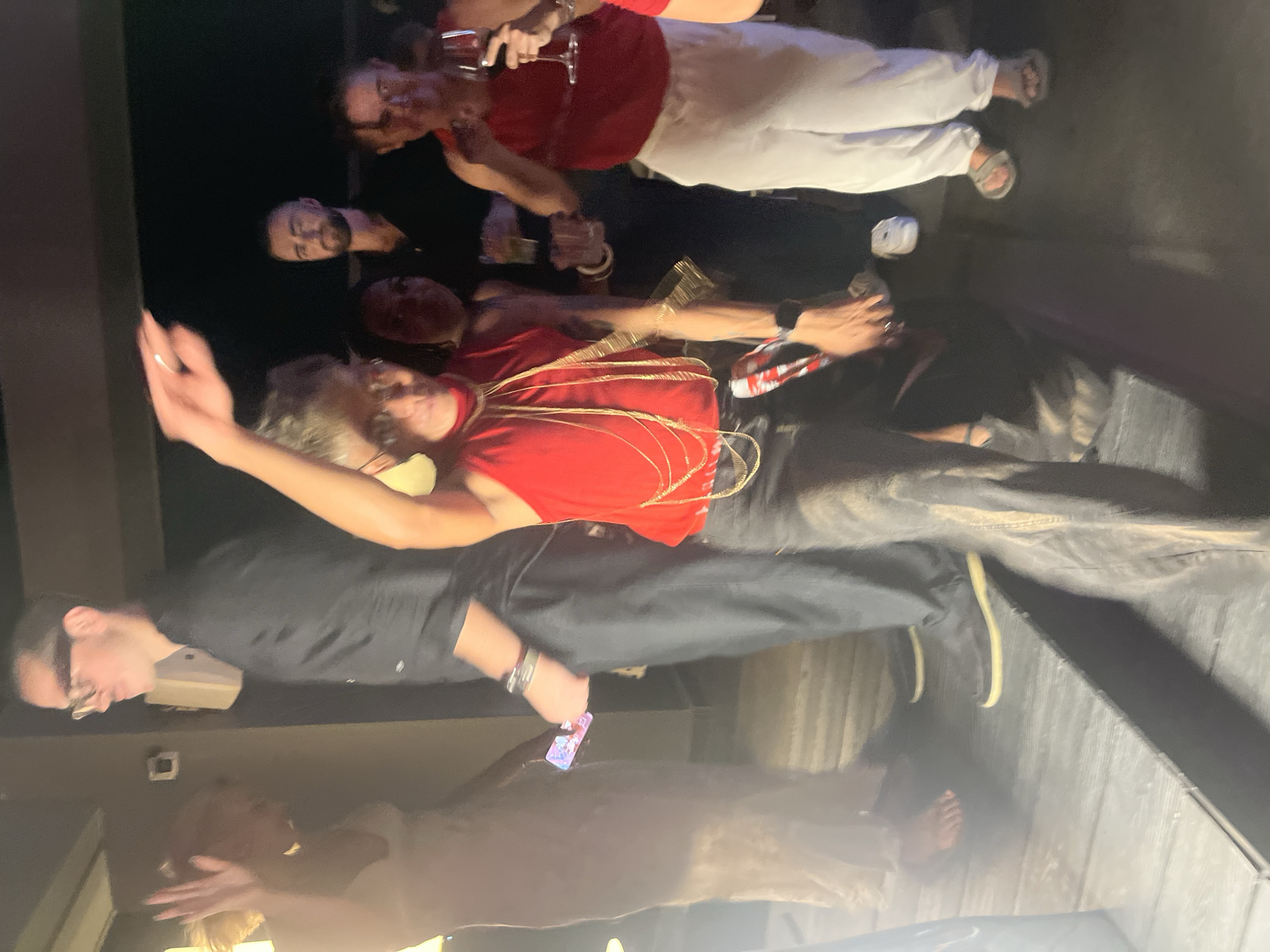 The image captures a group of people indoors, possibly at a bar or a private party, with a rustic wooden floor. The lighting is dim, suggesting it's nighttime. Several individuals are visible, some in the foreground and others in the background. In the foreground, a woman in a red top is holding a glass and appears to be dancing or posing. She has a decorative gold rope draped around her. Beside her, a man in a black shirt and glasses is looking down. Further back, another person is seen in a red top and white pants. There are other individuals in the background, some holding drinks. The overall atmosphere seems to be one of celebration or a social gathering. No text is visible in the image. The image is oriented sideways.