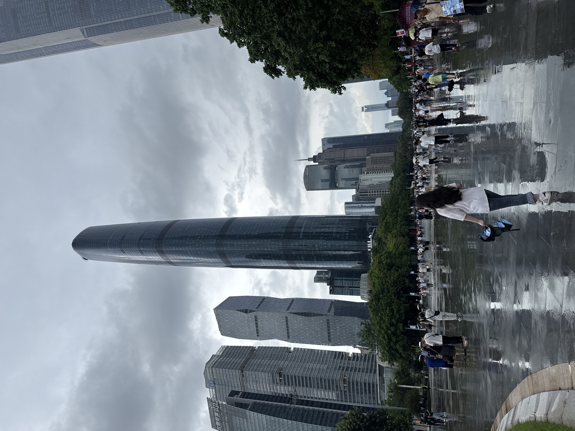 In Tianhe District, China, under an overcast sky, a wide, wet, paved plaza reflects the surrounding modern skyscrapers. The foreground shows the wet surface of the plaza with a woman in a white shirt and blue jeans walking away from the camera, holding a closed black umbrella. She is wearing white sneakers with black stripes. Further into the scene, numerous people are walking in various directions across the plaza. Some are holding umbrellas, indicating recent or ongoing rain. The architecture is dominated by tall, sleek glass and steel buildings, with one particularly tall, dark, cylindrical skyscraper standing prominently in the mid-ground. Green trees line the edges of the plaza and are interspersed between some of the buildings. The overall atmosphere is one of urban activity after a period of rain.