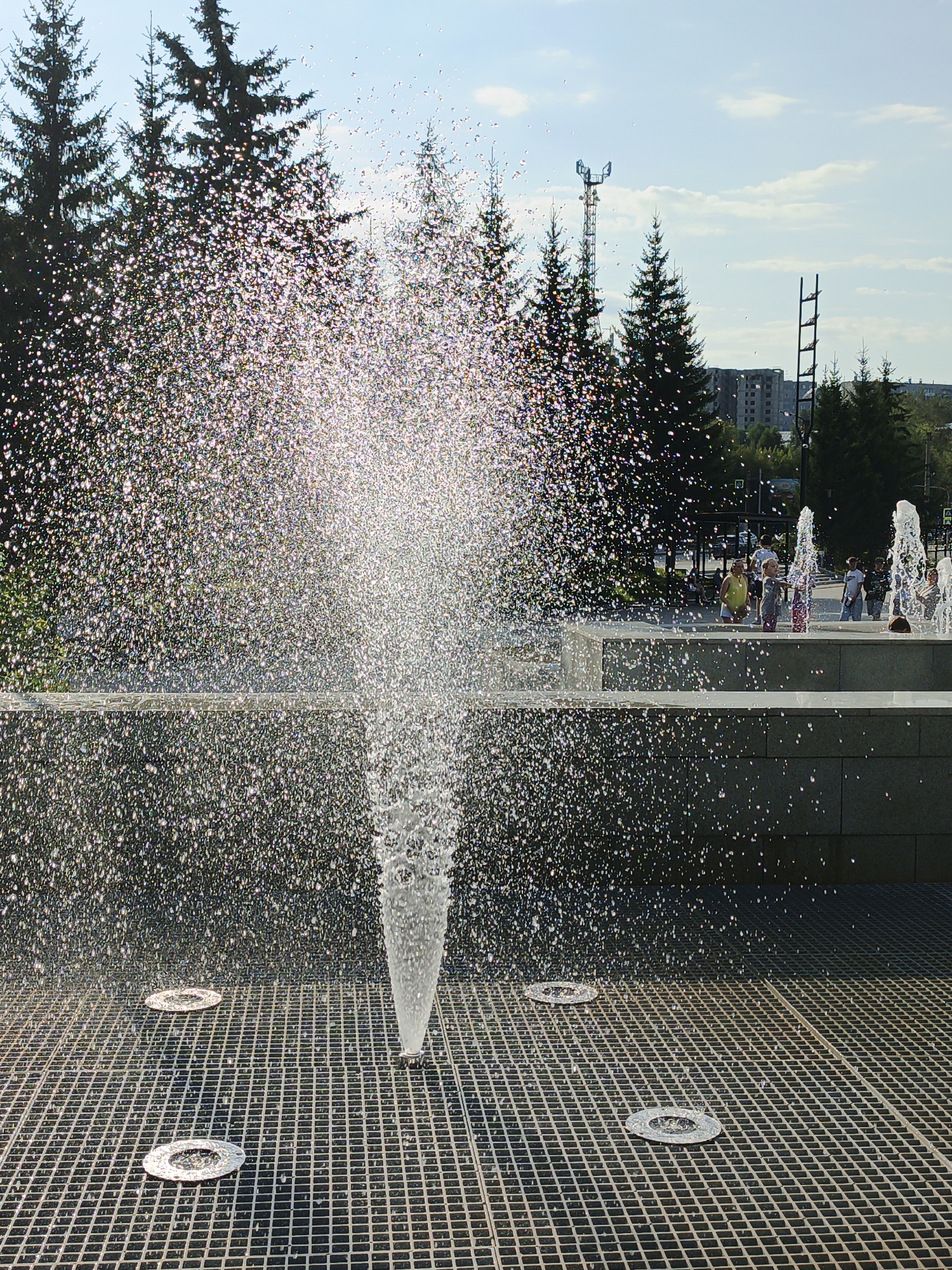 This image depicts a public fountain on a sunny day in the city of Zlatoust, Russia. The foreground features a metallic grate with circular openings, some of which appear to be water jets. A central jet of water is spraying upwards in a cone shape, creating a misty effect that refracts the sunlight into a spectrum of colors. In the middle ground, there is a tiered concrete structure with several more water jets active. A group of children and adults are visible on this tiered level, with some children playing in or near the water. Behind the fountain, a line of tall evergreen trees forms a natural backdrop. In the distance, a telecommunications tower and some low-rise buildings, including a partially constructed one, are visible under a partly cloudy blue sky. The overall scene suggests a recreational area in an urban park or plaza.
